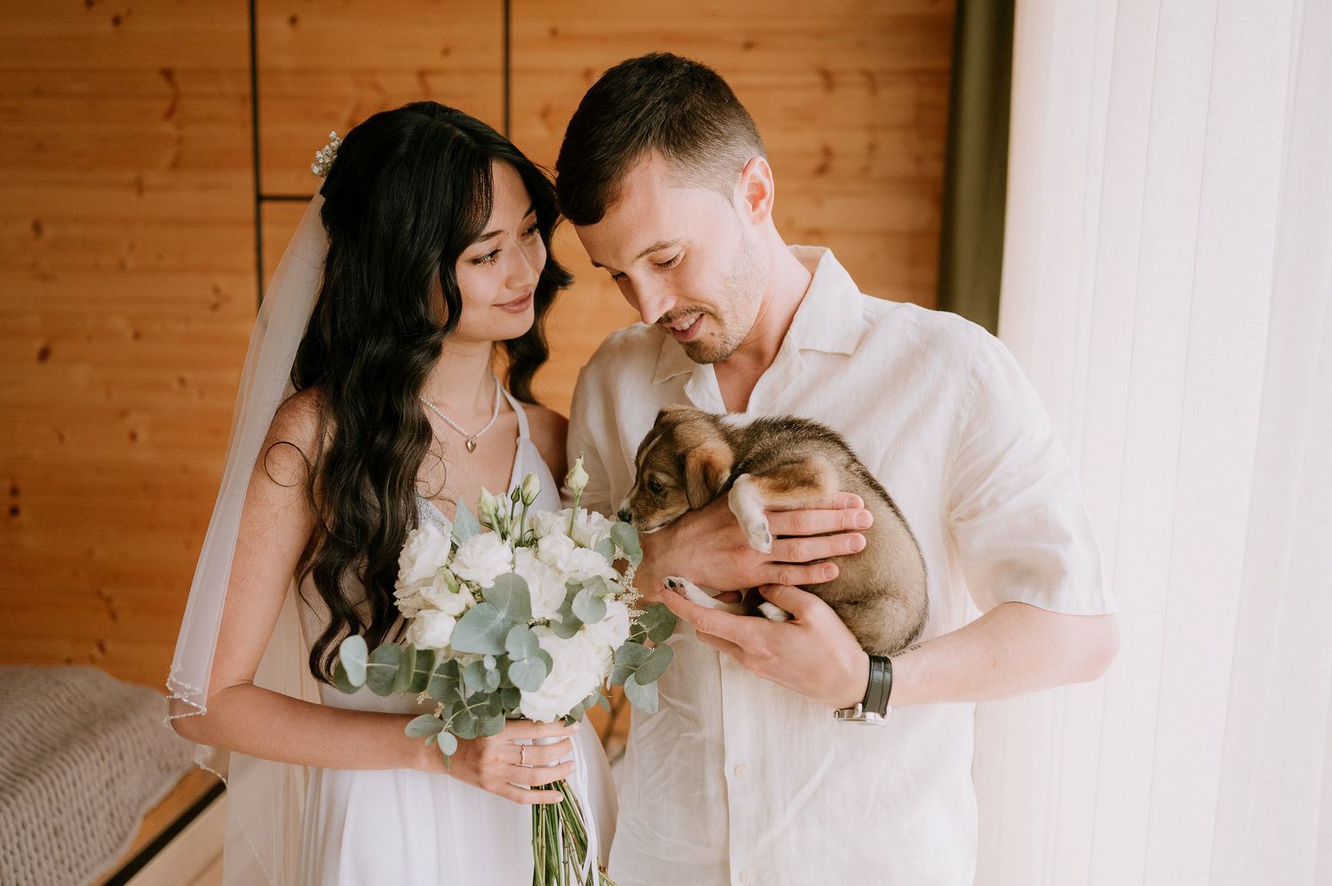 A couple in white embraces while holding a bouquet of white flowers with eucalyptus in a wooden interior.
