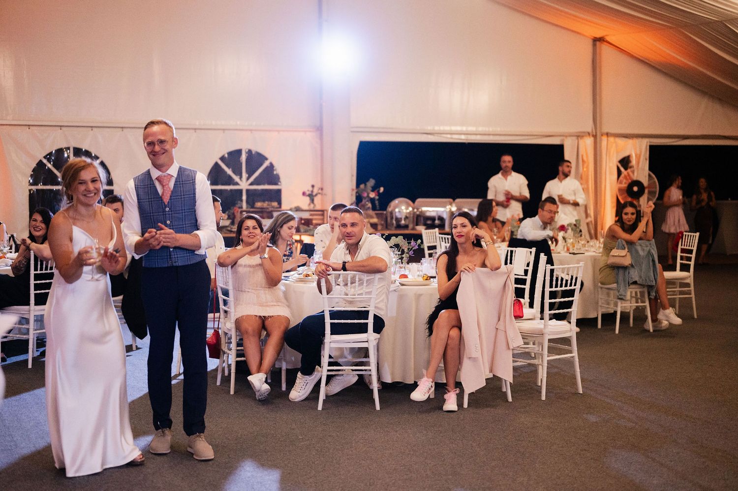 Wedding party dancing and celebrating in a decorated reception tent with white chairs.