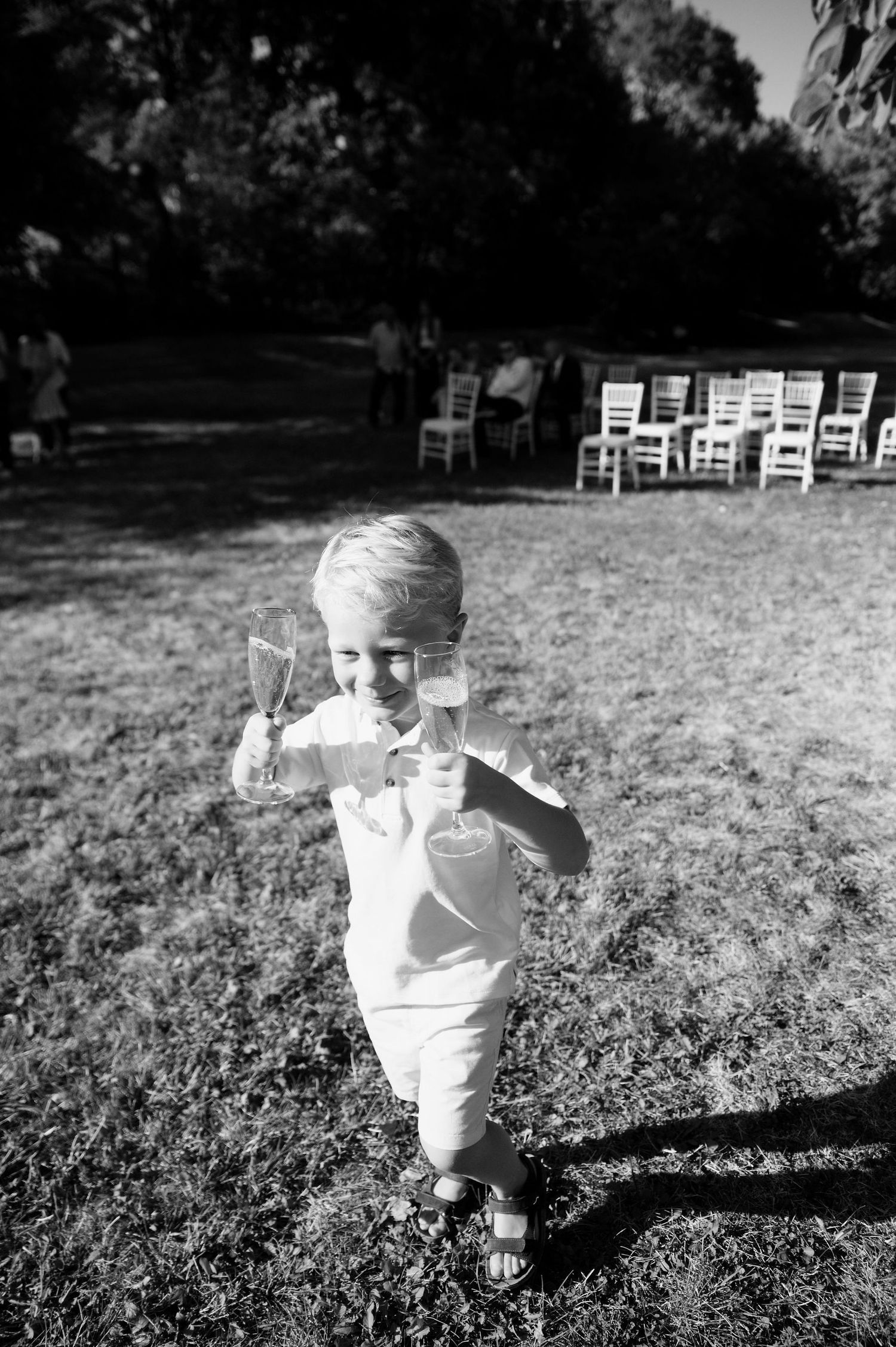A small child in white clothing runs across a grassy field with chairs in the background.