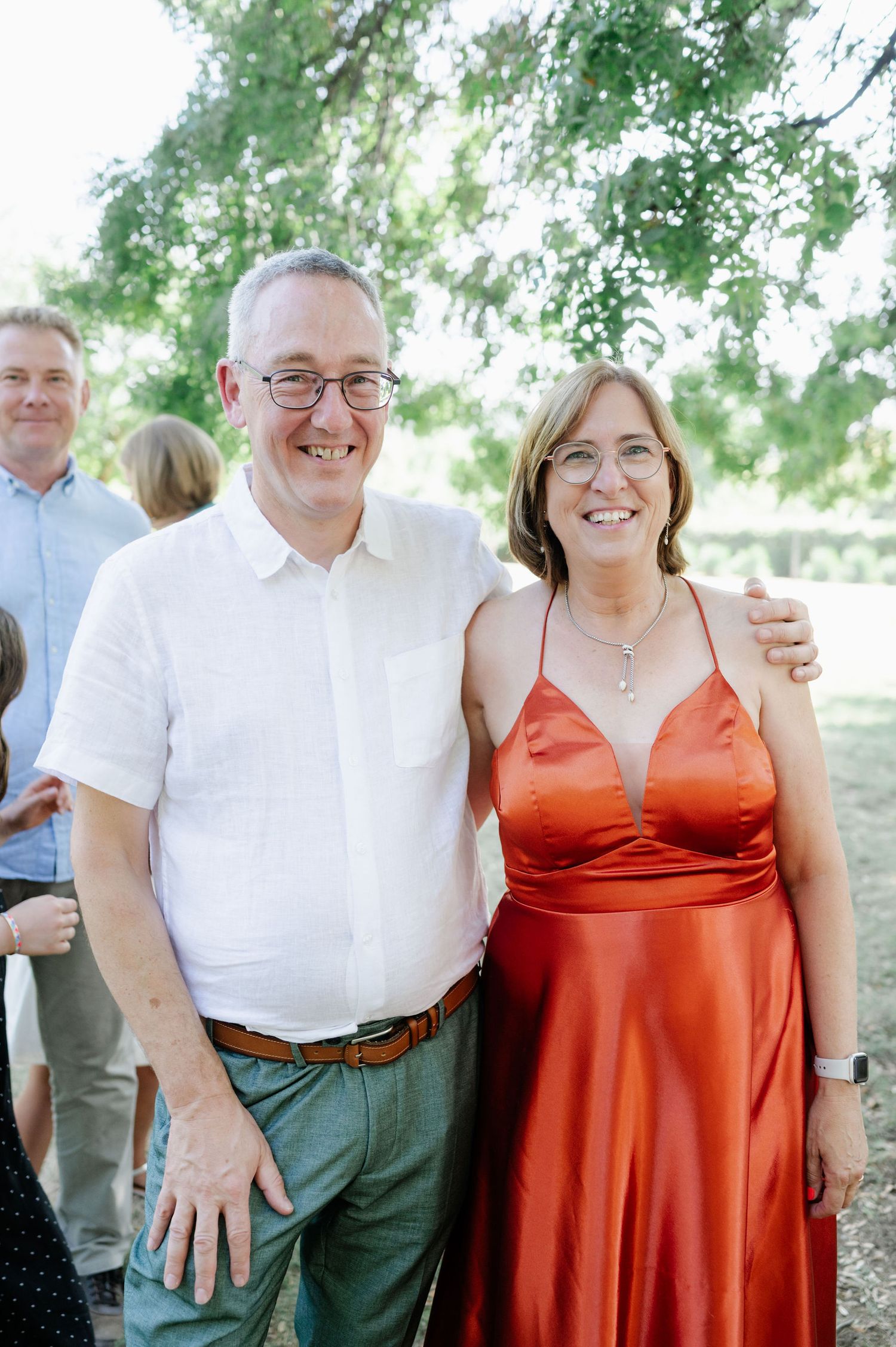 Couple in formal attire posing at an outdoor gathering, wearing white and orange.