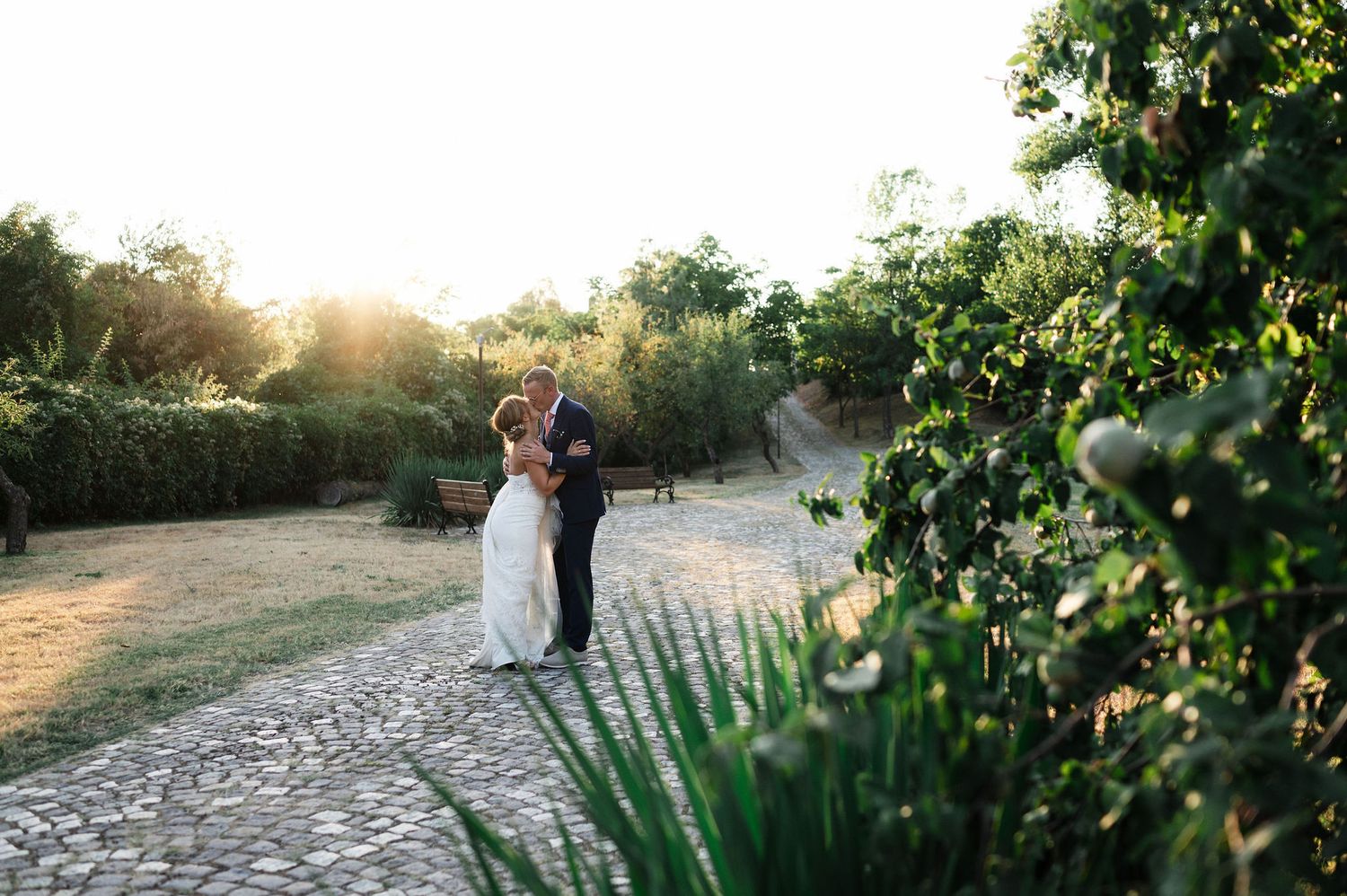 Distant shot of newlyweds on a garden path with golden sunlight filtering through trees.
