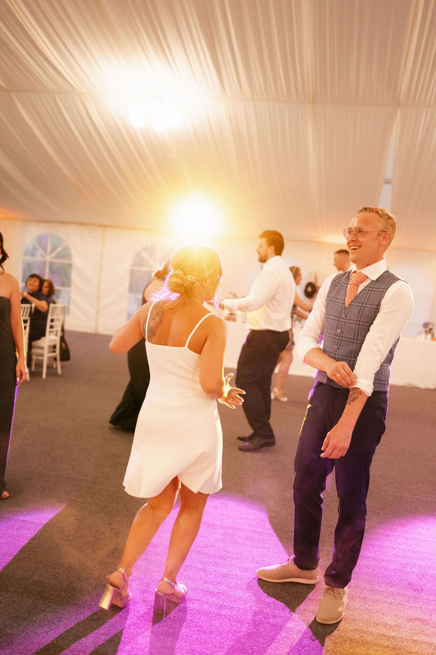 Wedding guests dance and socialize under tent lighting with purple floor accents.