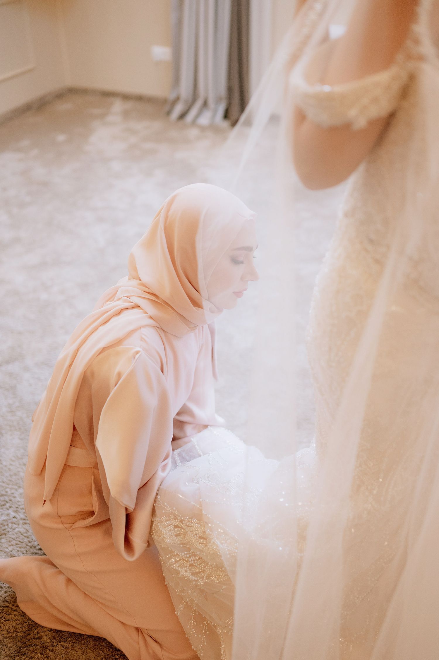 Person in light pink hijab kneeling beside flowing white wedding dress.