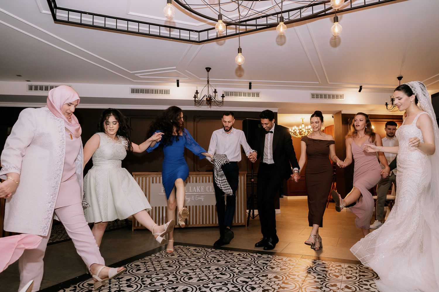 Wedding guests dance and celebrate on a black and white patterned floor under pendant lights.
