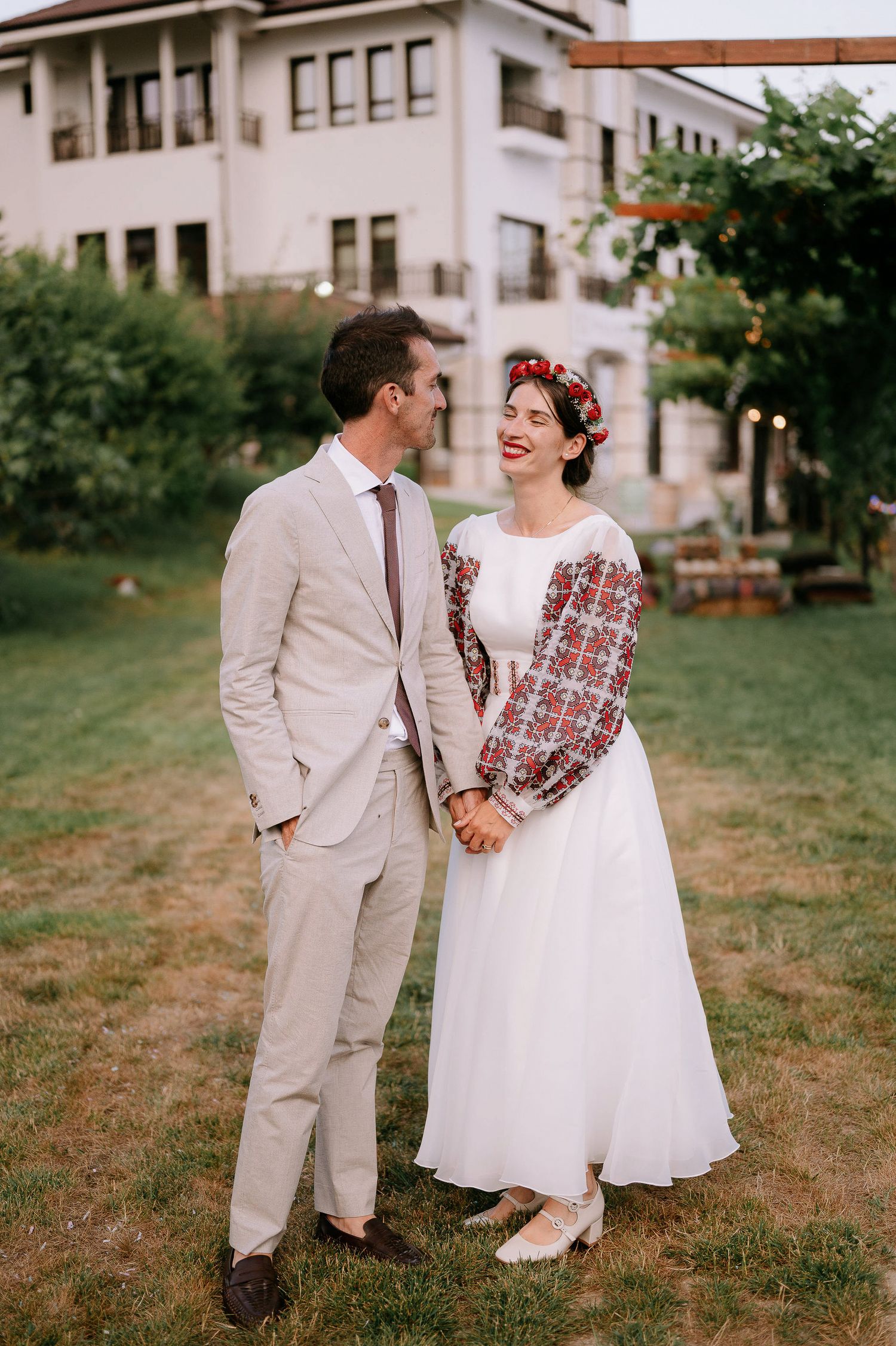 A joyful wedding couple laughing together on a grassy slope wearing coordinated vintage-inspired outfits.