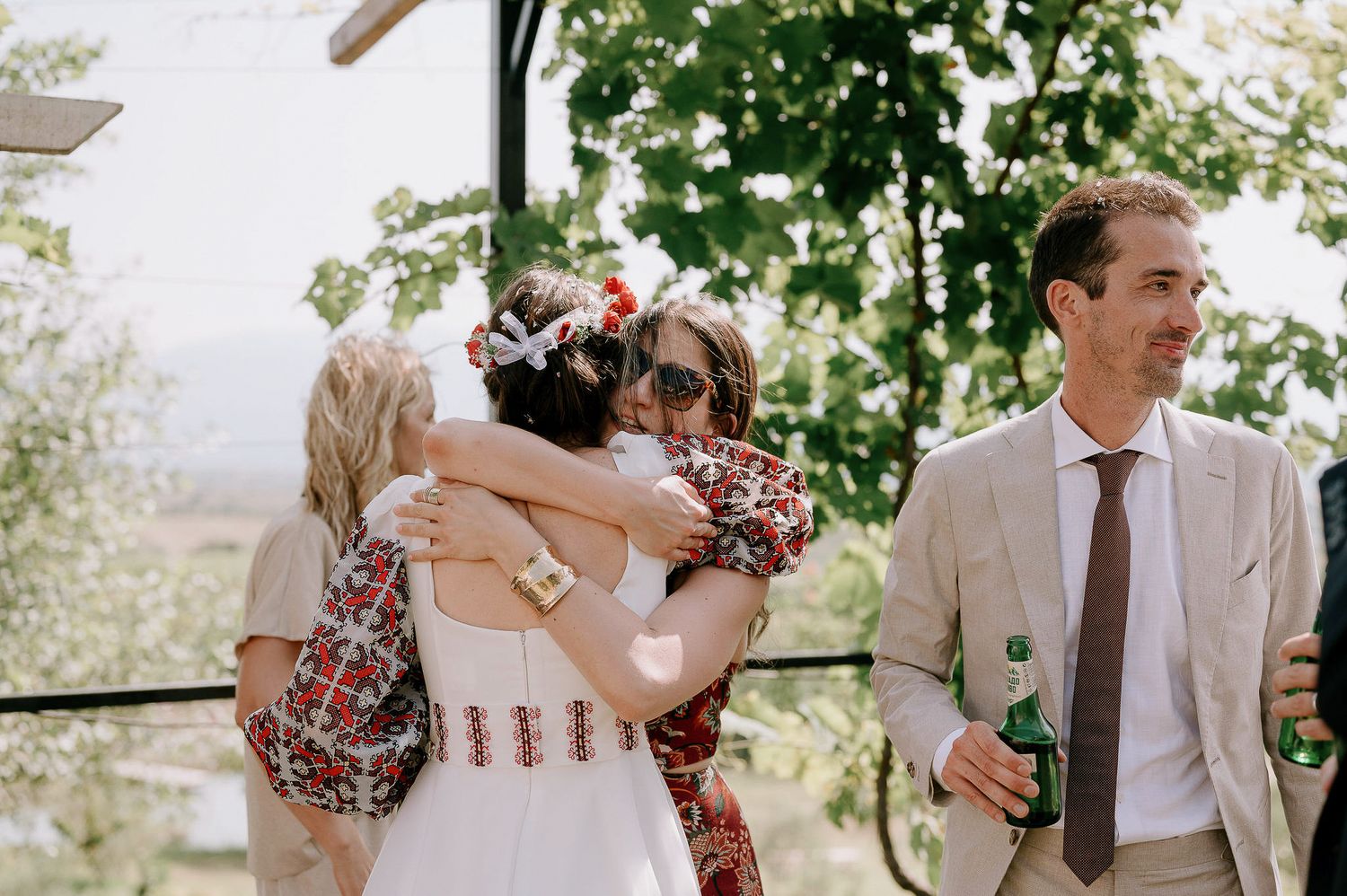 Wedding guests gather outdoors under trees at a summer celebration.