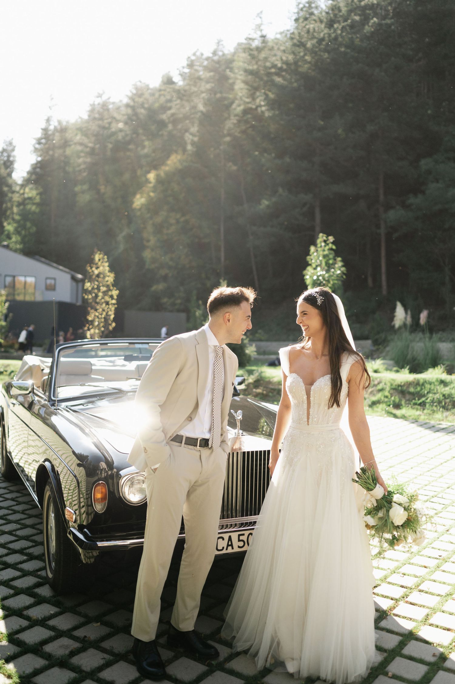 Sunlit wedding portrait with a classic black Rolls Royce on decorative stone pavement.
