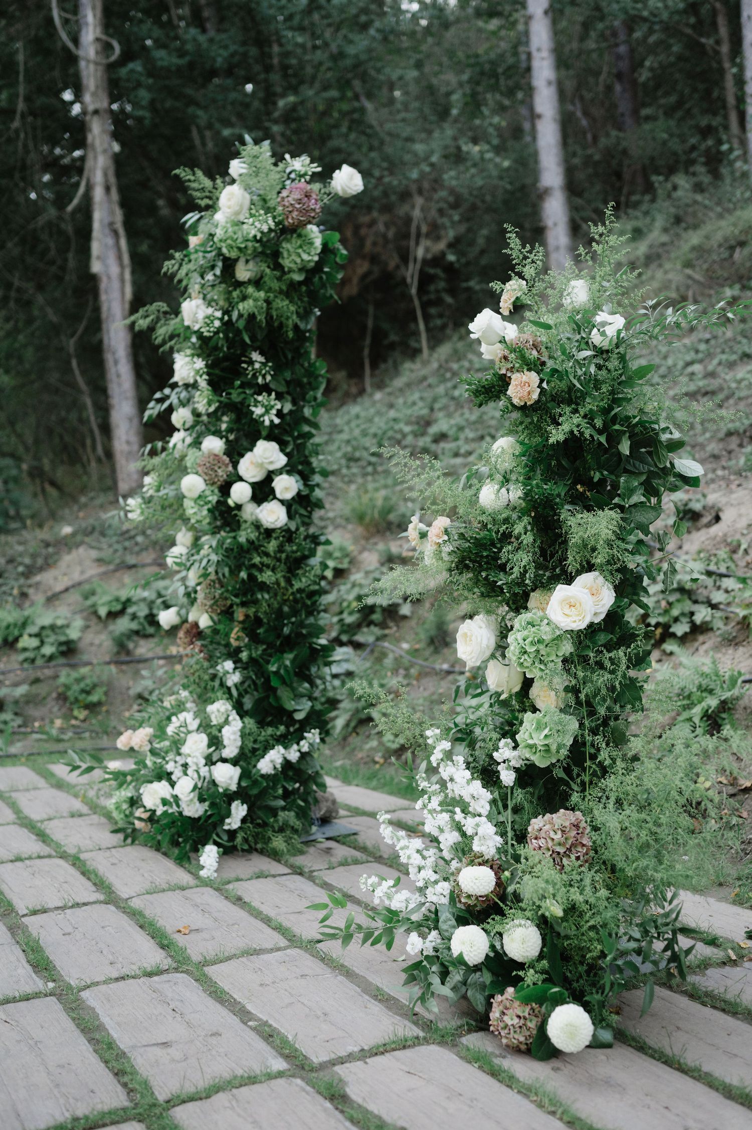 Stone pathway lined with lush white rose arrangements and greenery in a woodland garden setting.