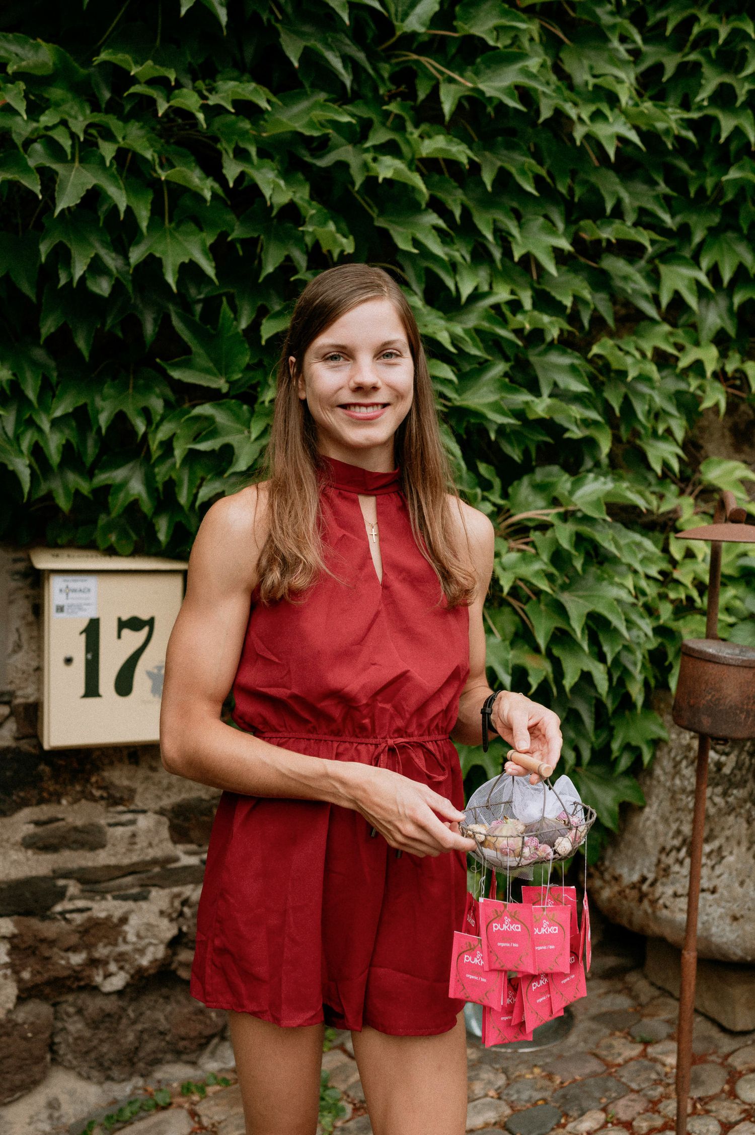 A woman in a red romper stands against an ivy-covered wall holding a small gift bag.