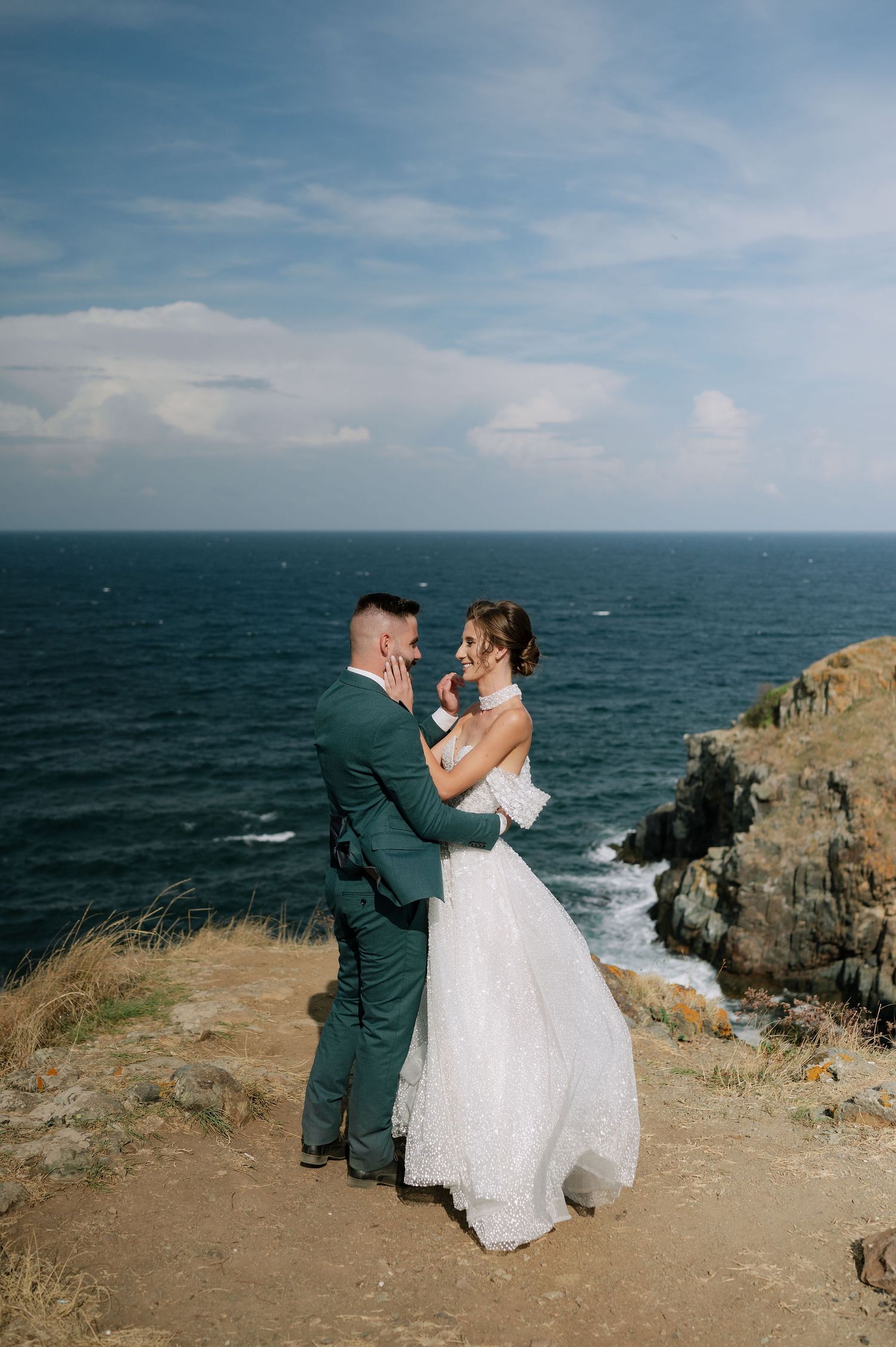 A wedding couple poses dramatically on a seaside cliff with waves crashing below.