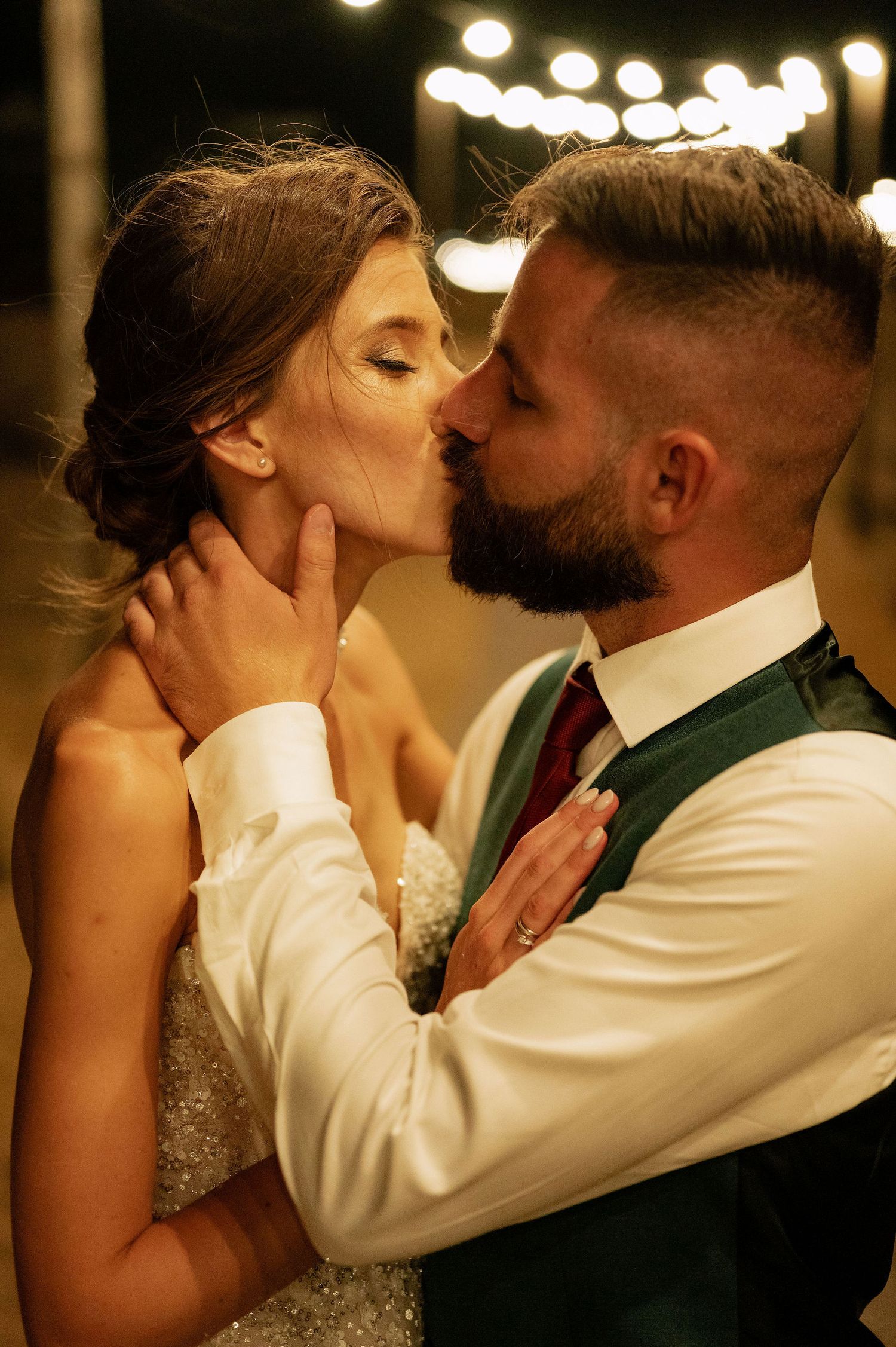 An intimate kiss shared between newlyweds under romantic string light illumination.