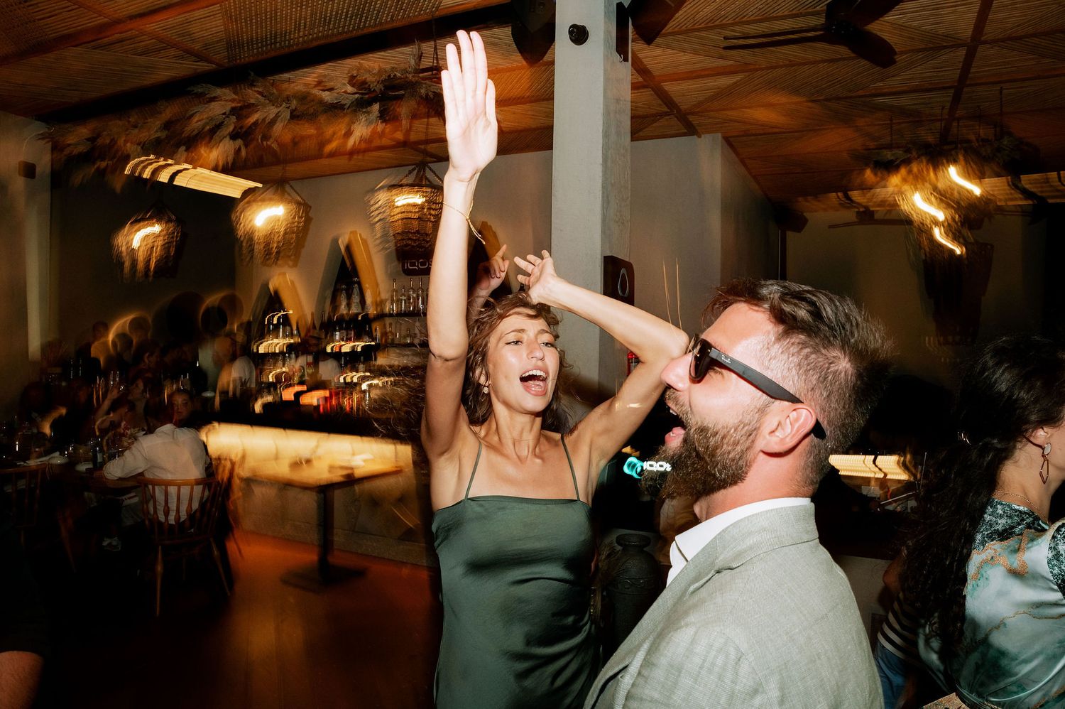 Wedding guests dancing enthusiastically with raised arms under warm lighting at an indoor reception.