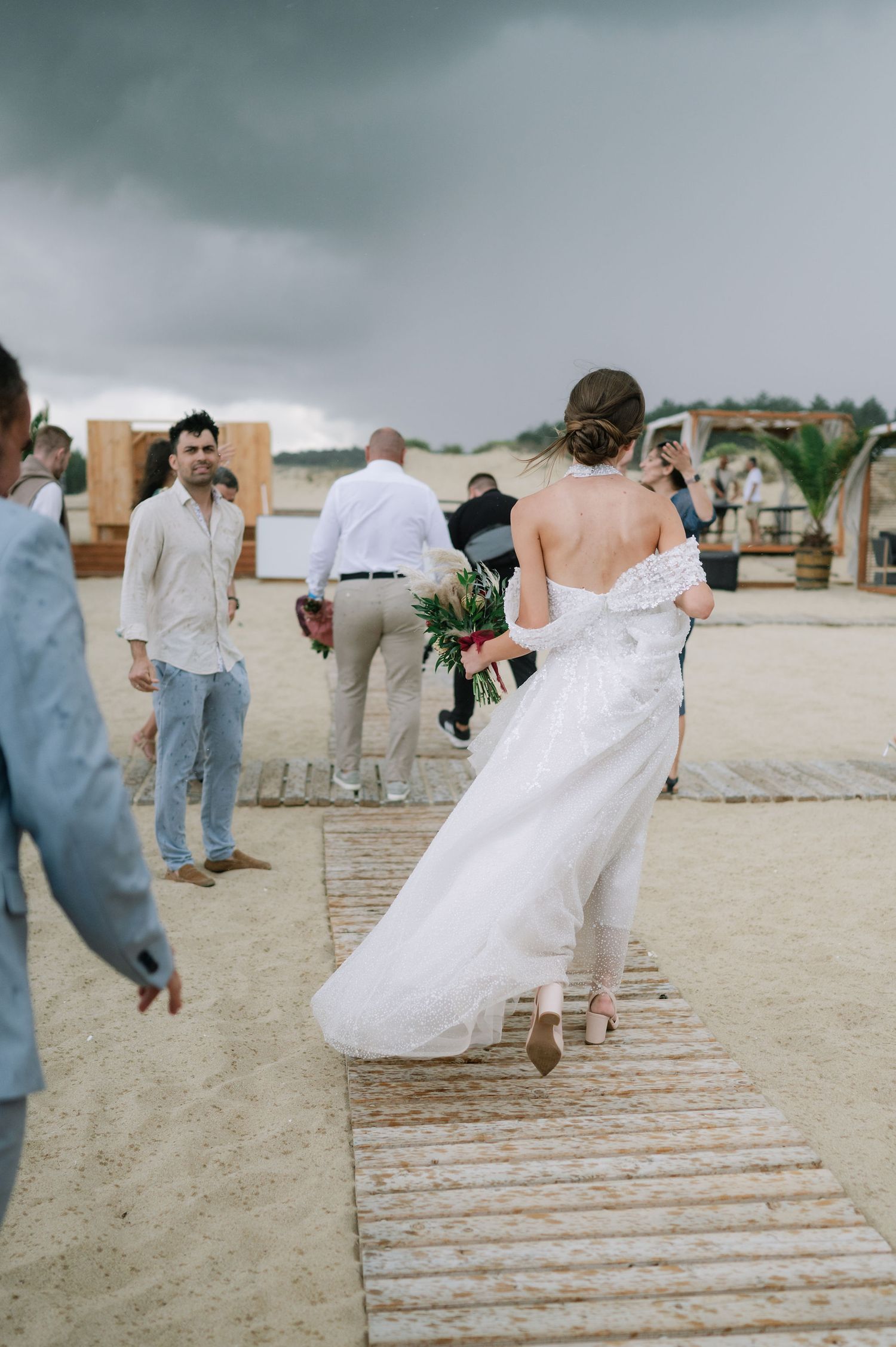 A couple walks down a wooden boardwalk on a beach during their wedding celebration.