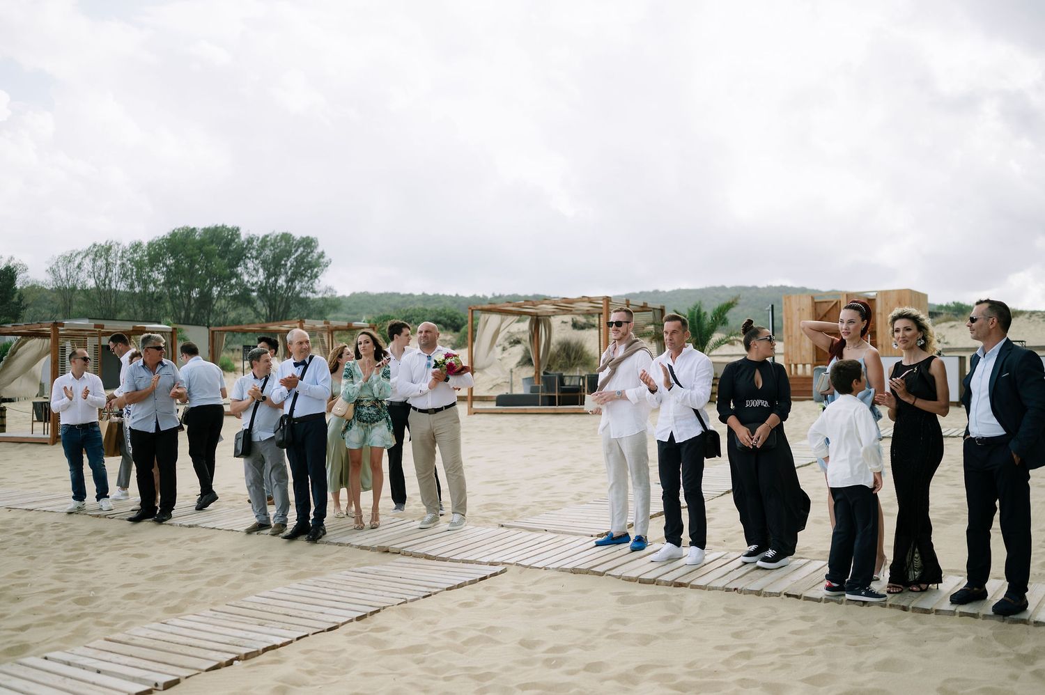Large group of wedding guests lined up on a wooden boardwalk by the ocean.