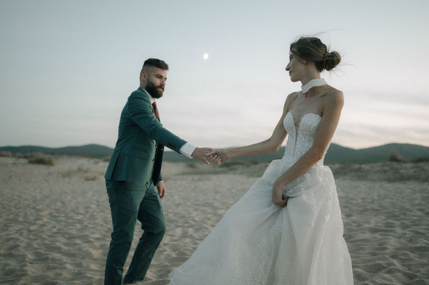A couple in wedding attire holds hands on a sandy beach at dusk, with a full moon visible in the twilight sky.