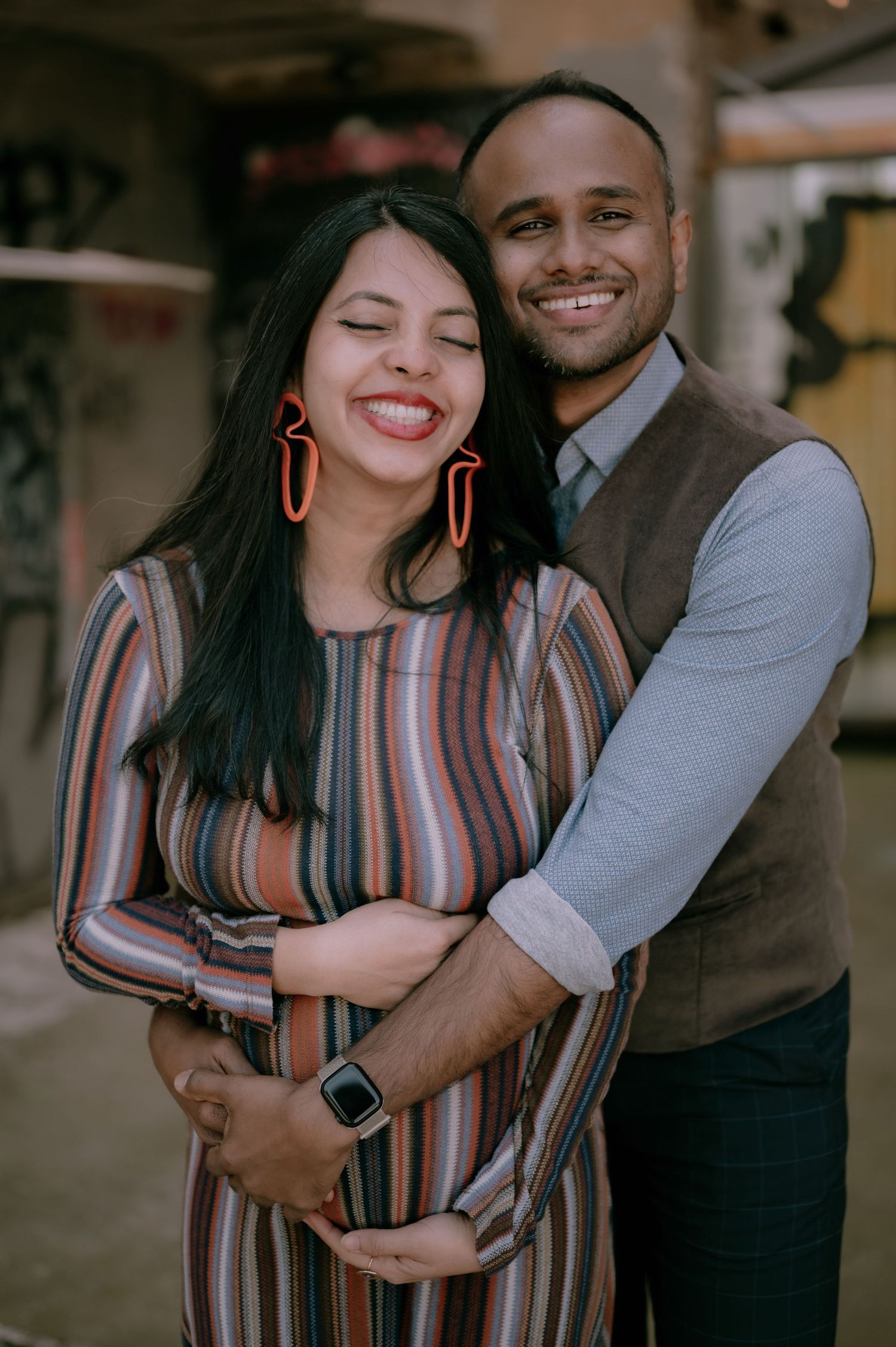 A smiling couple embraces tenderly, the woman in a striped dress with red earrings, the man in a vest, in a maternity photo.