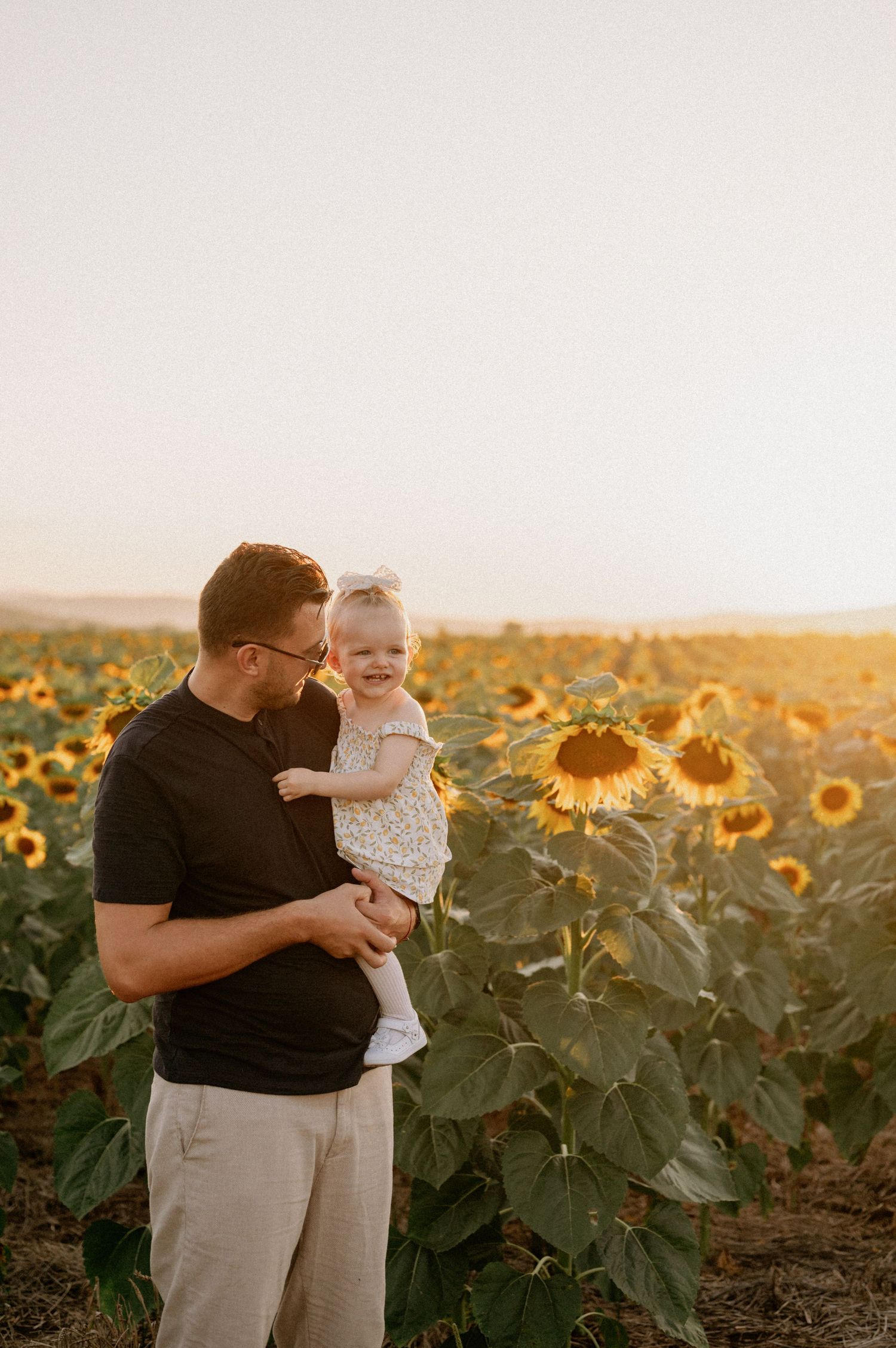 A father holds his laughing toddler in a golden sunflower field at sunset.