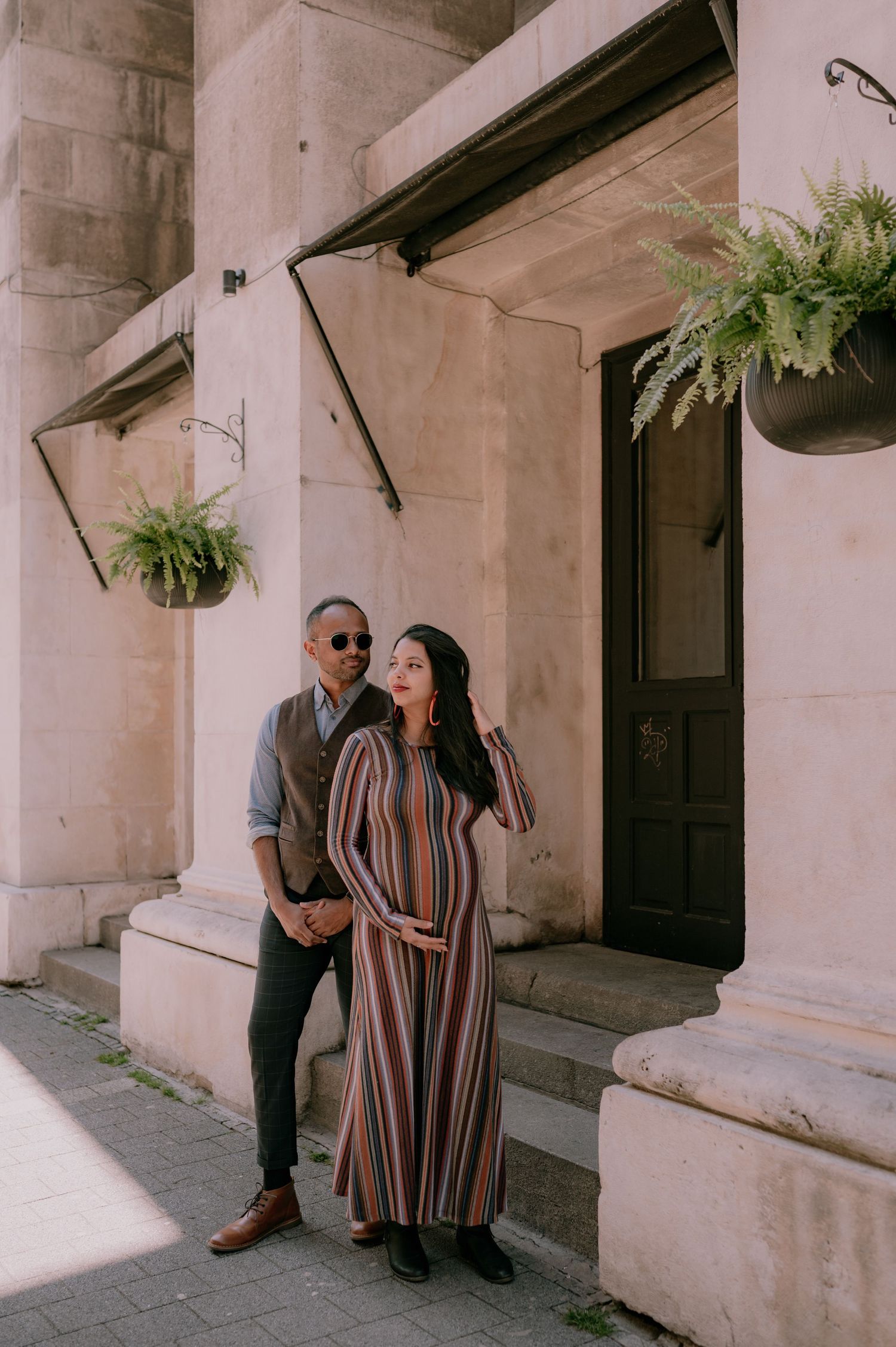 Pregnant couple posing outside a stone building with hanging ferns, woman in striped maxi dress, man in vest and sunglasses.