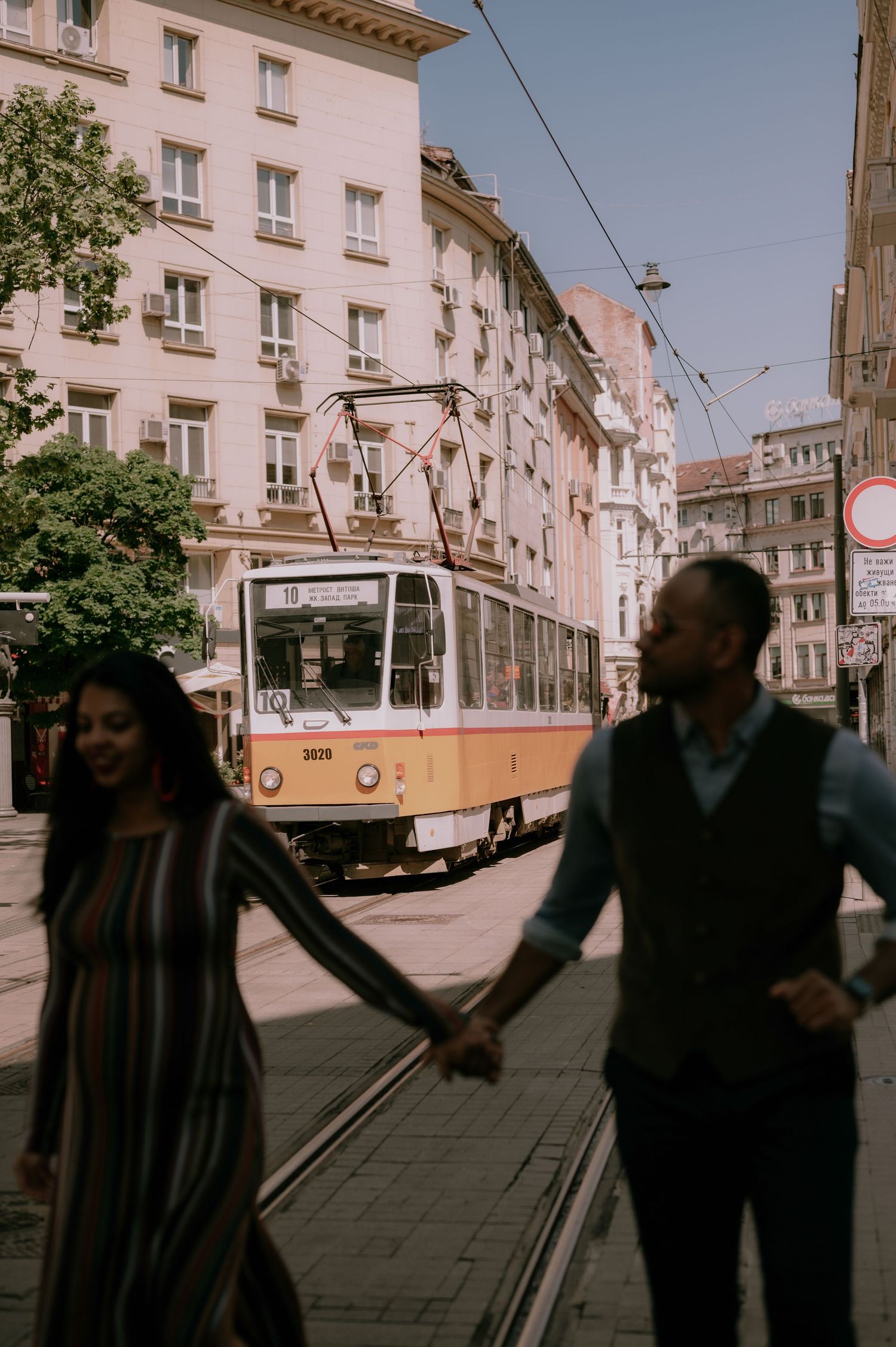 Couple holding hands crossing a city street with a vintage yellow tram and European buildings in the background.