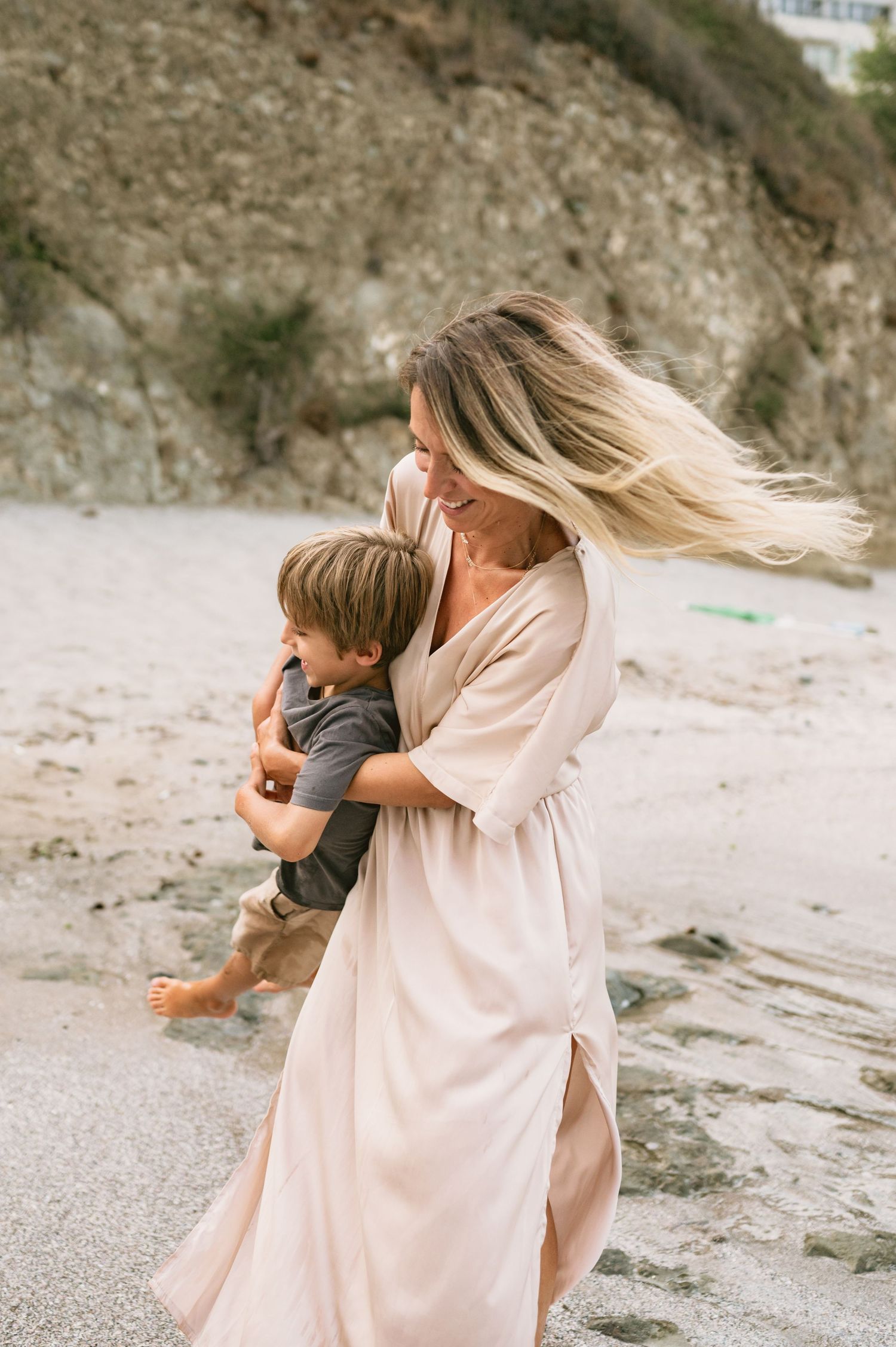 A woman in a flowing blush dress hugs a young boy on a sandy beach with rocky cliffs in the background.