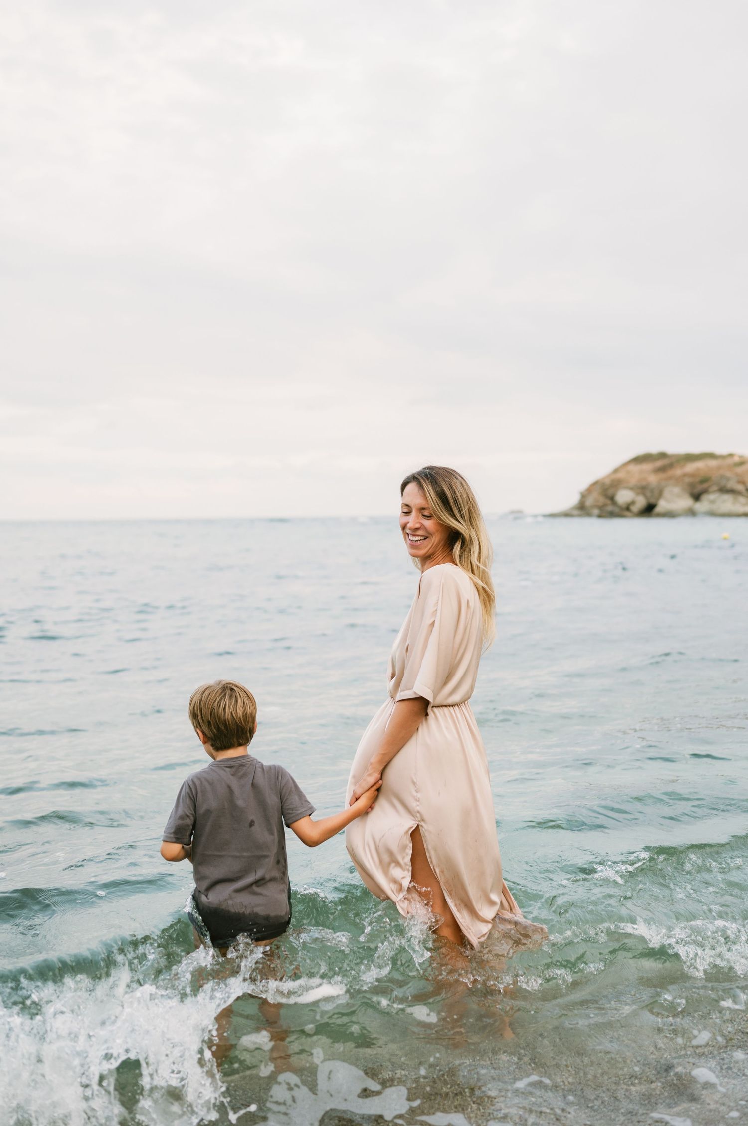 A smiling woman in a pink dress holds hands with a young boy as they wade together in ocean waves.