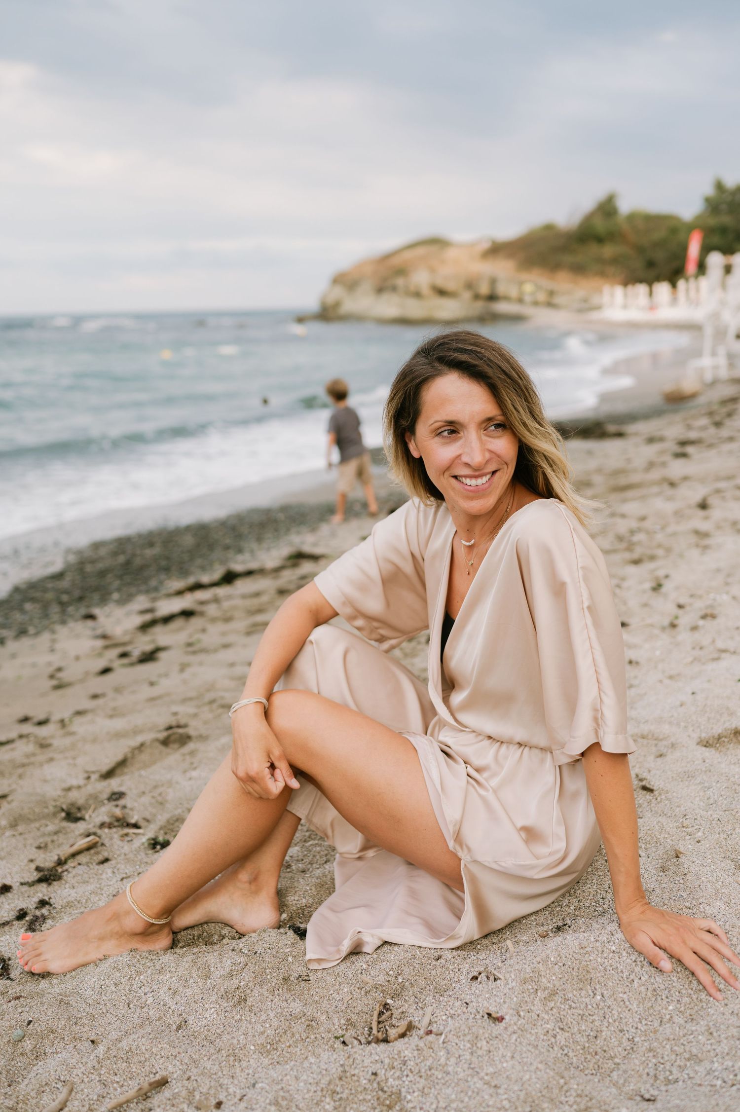 Woman in beige dress sitting on sandy beach smiling, with ocean waves and rocky headland in background.