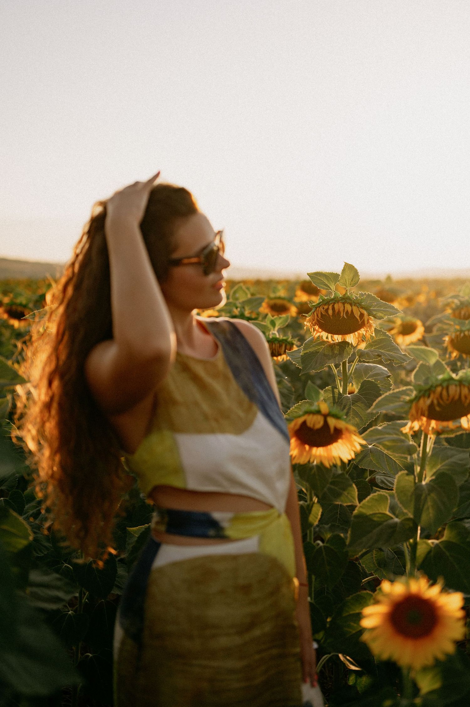 Woman in sunglasses posing in a sunflower field during golden hour sunset light.