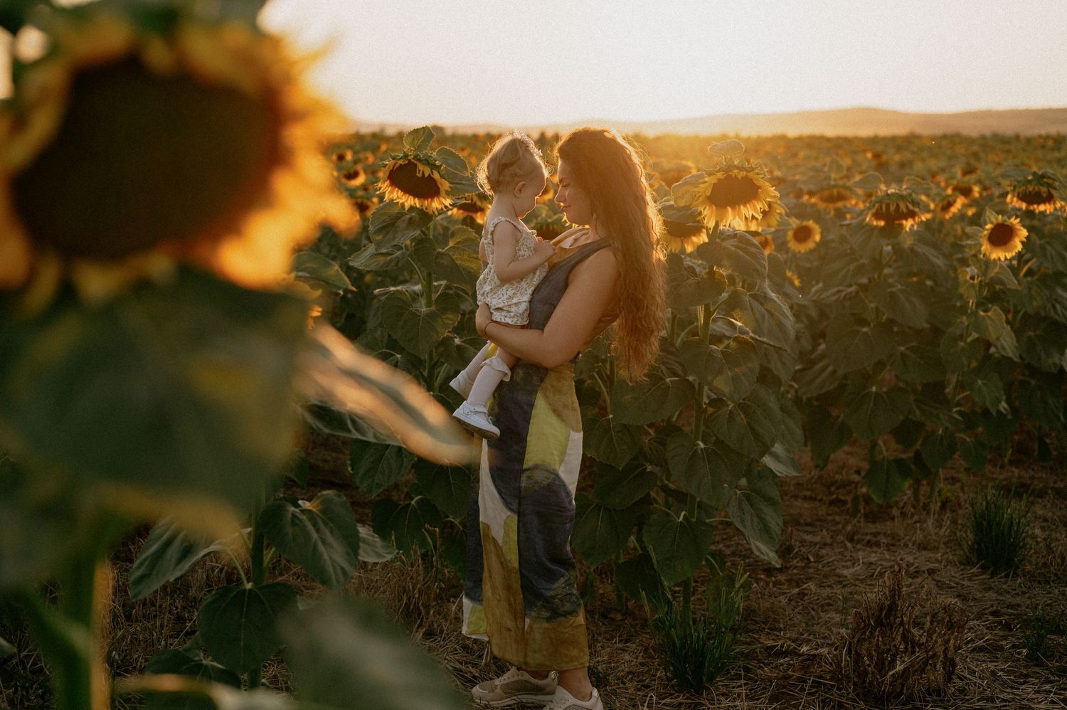 A mother lifts her toddler in a golden sunflower field at sunset, sharing a tender moment.