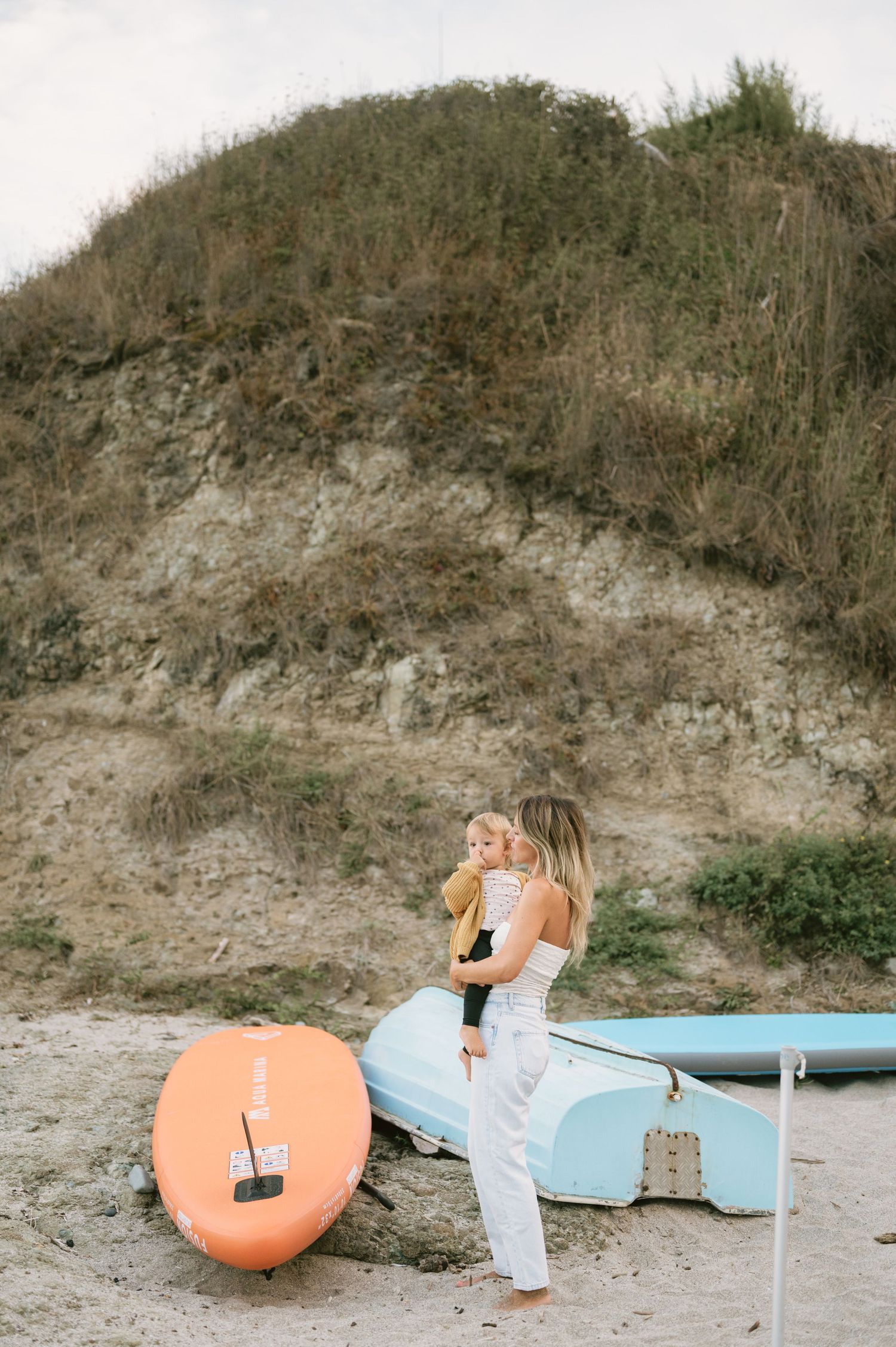Woman holding toddler on sandy beach beside orange paddleboard and blue boat with grassy dune backdrop.