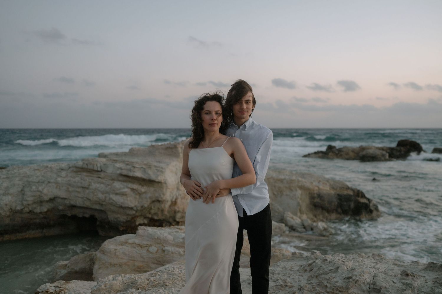 Couple embracing on rocky coastal cliffs at sunset with ocean waves in background.