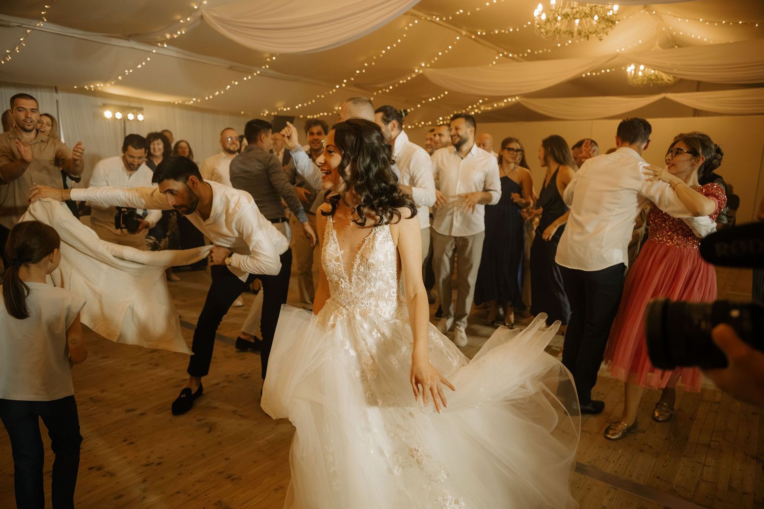 Bride in white gown dancing joyfully with wedding guests on a decorated reception dance floor.