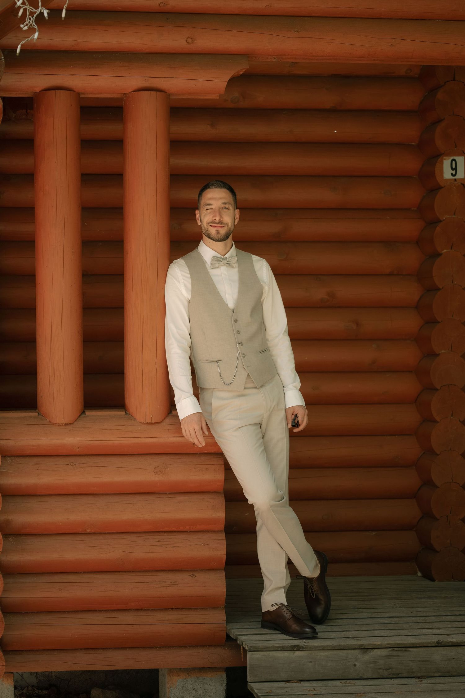 Smiling groom in beige vest and bow tie leaning against wooden log cabin porch column.