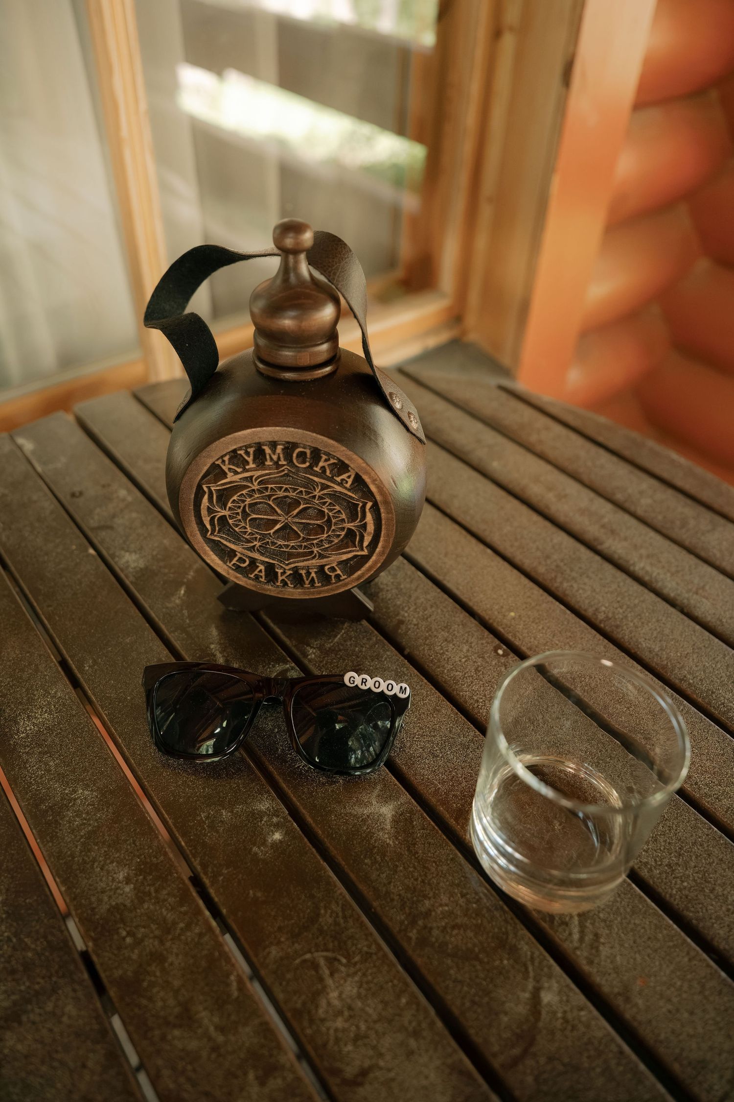 Rustic wooden table with a decorative Kymoka Parks flask, sunglasses, and a glass near a window.