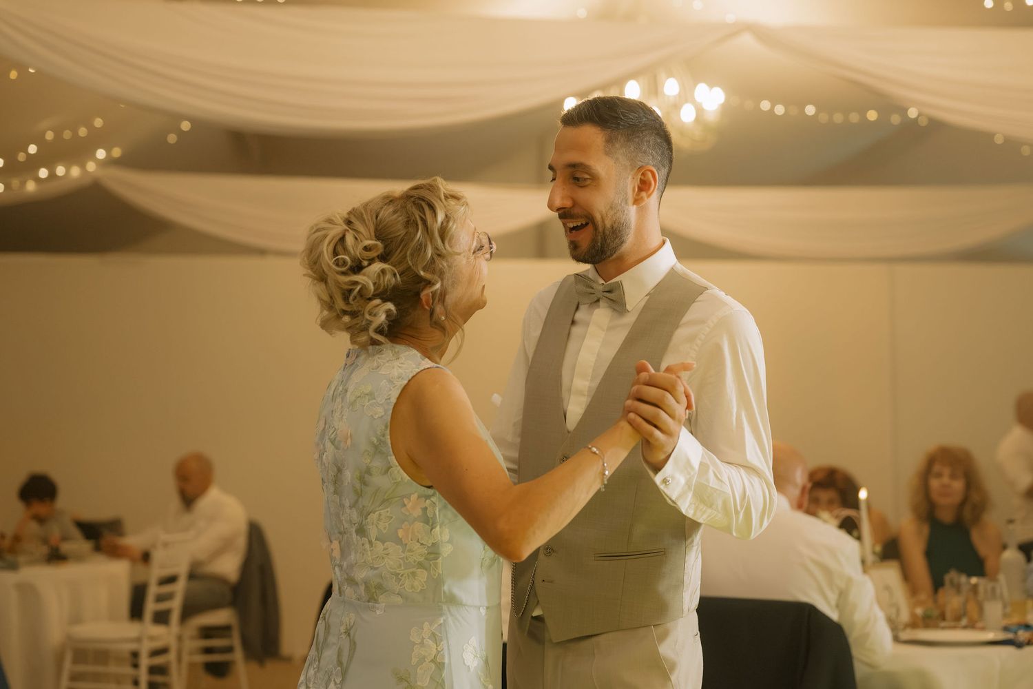 Groom in gray suit shares first dance with woman in blue dress at elegant wedding reception with fairy lights.