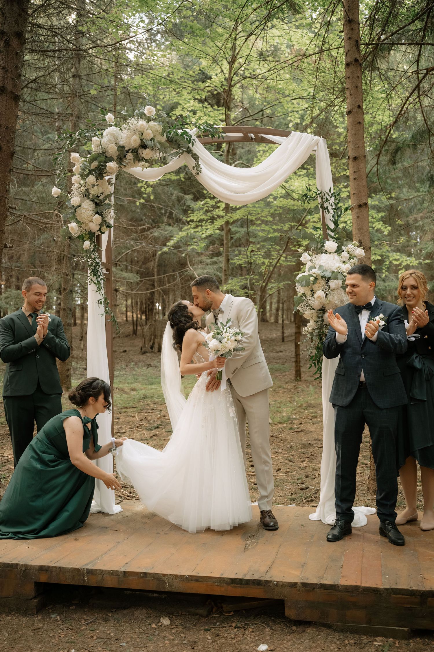 Bride and groom share first kiss at outdoor forest wedding ceremony with floral arch and guests applauding.