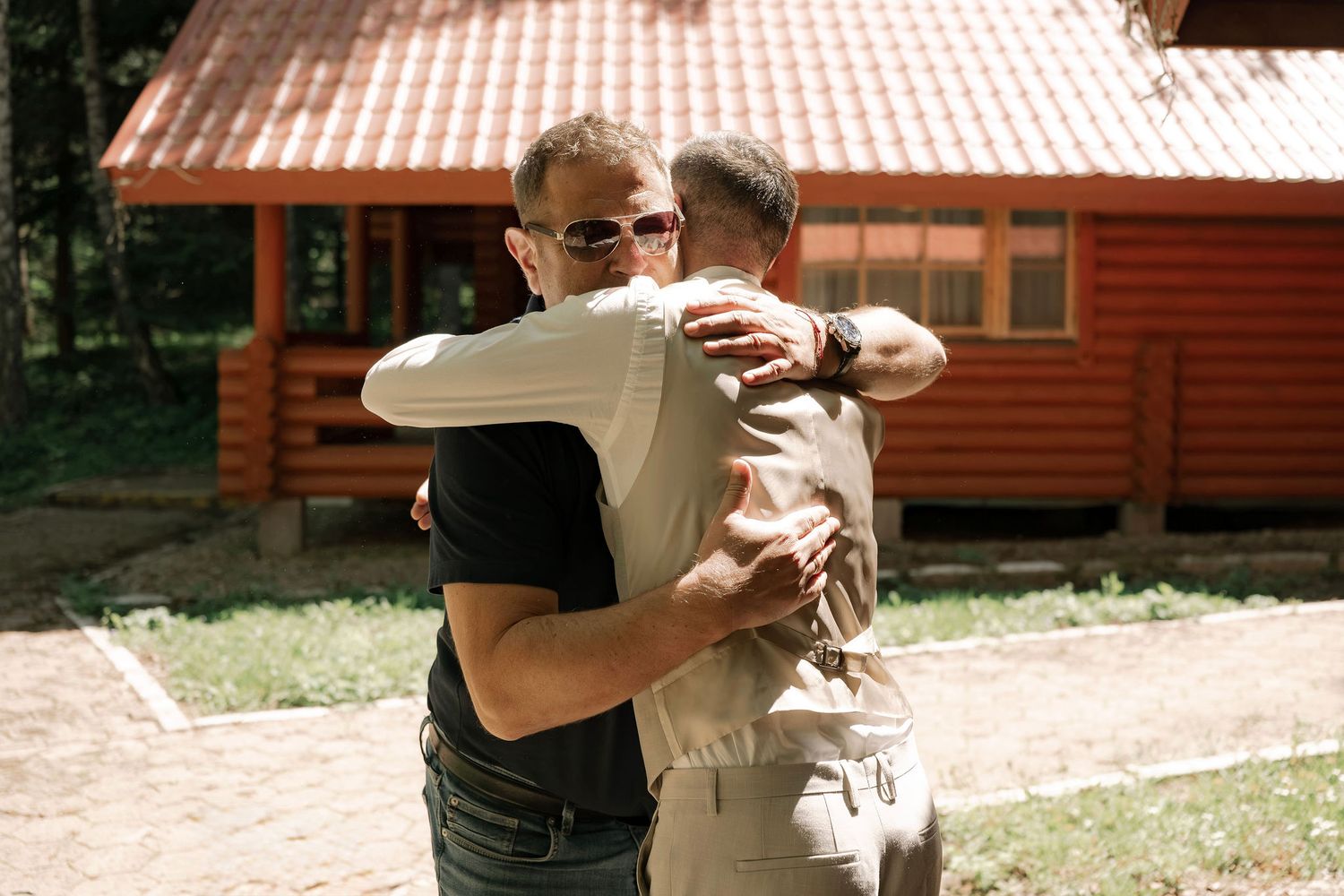 Two men sharing a warm embrace outdoors near a rustic log cabin surrounded by trees.