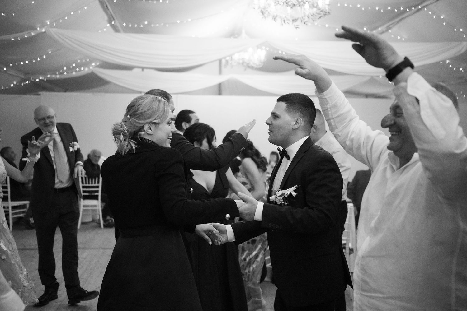 Black and white photo of guests dancing joyfully at an elegant wedding reception in a decorated venue.