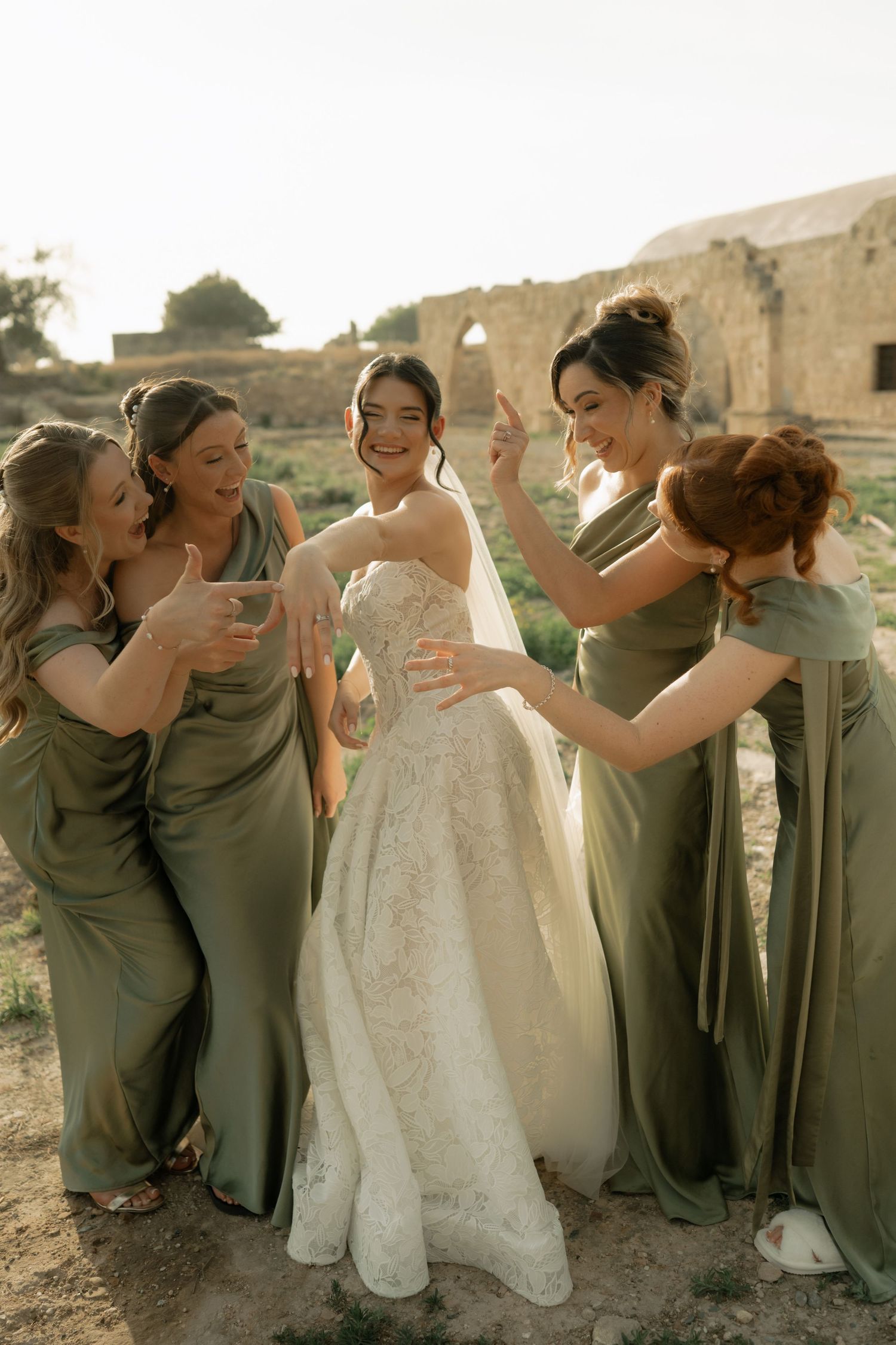 Bride in lace gown surrounded by smiling bridesmaids in sage green dresses at an outdoor rustic wedding venue.