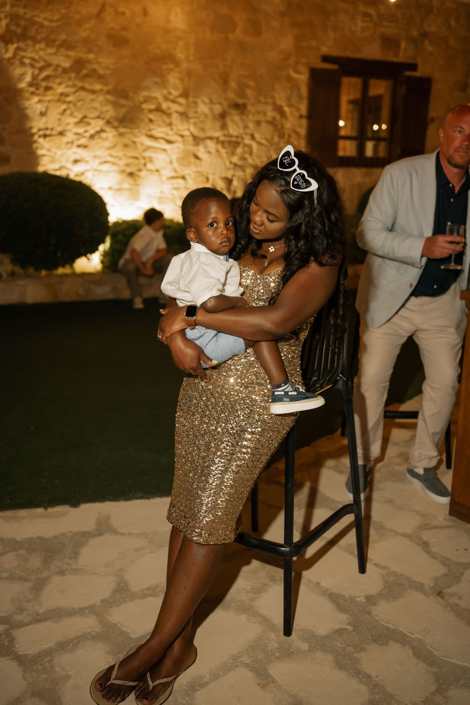 A woman in a gold sequin dress holds a baby at an elegant outdoor evening event with stone walls.