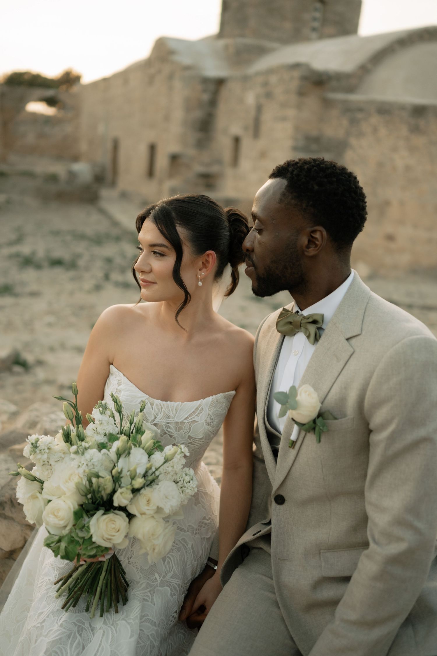 Bride holding white bouquet and groom in beige suit share an intimate moment near historic stone ruins.