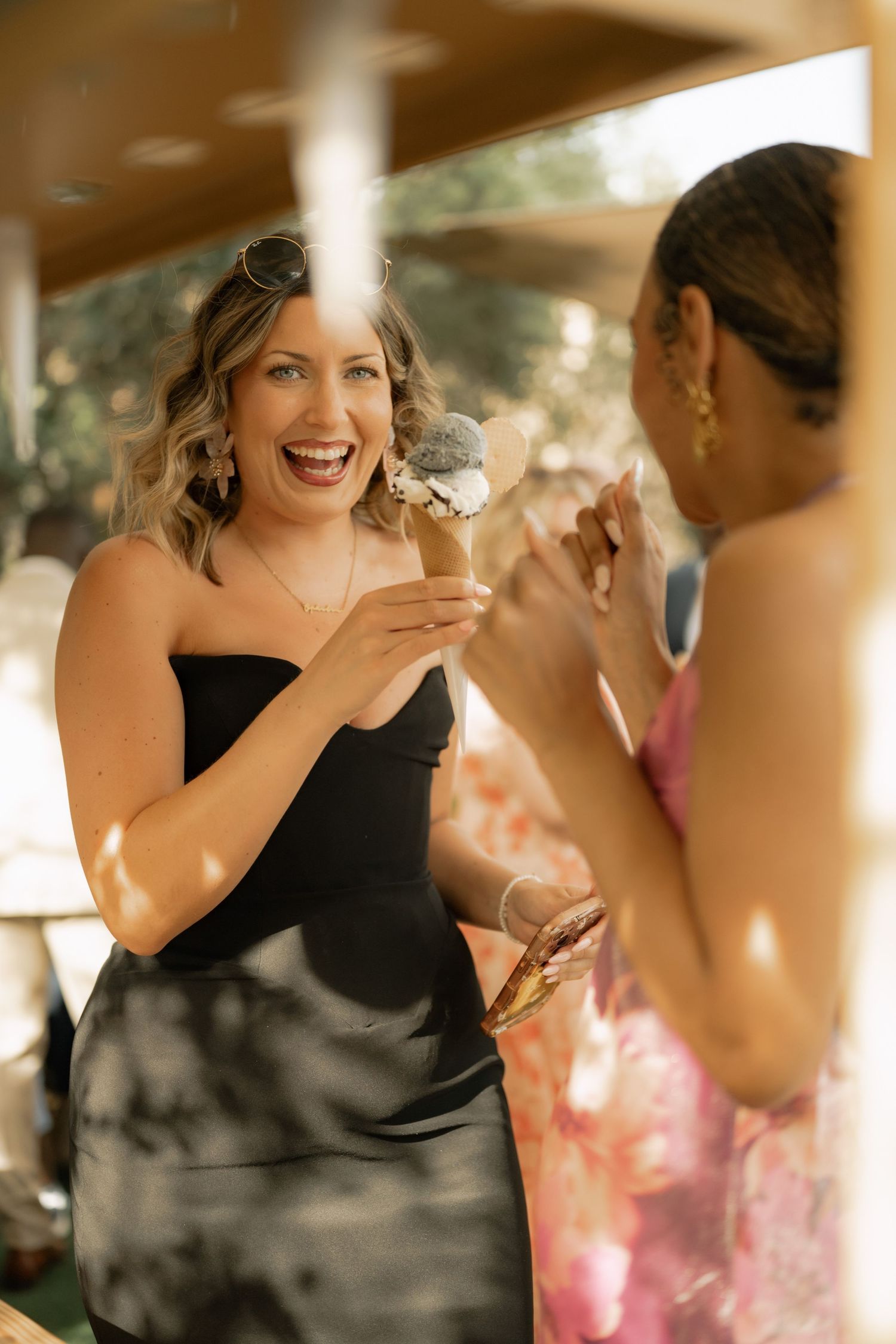 Two women laughing and enjoying ice cream cones outdoors on a sunny day, one wearing a black strapless dress.