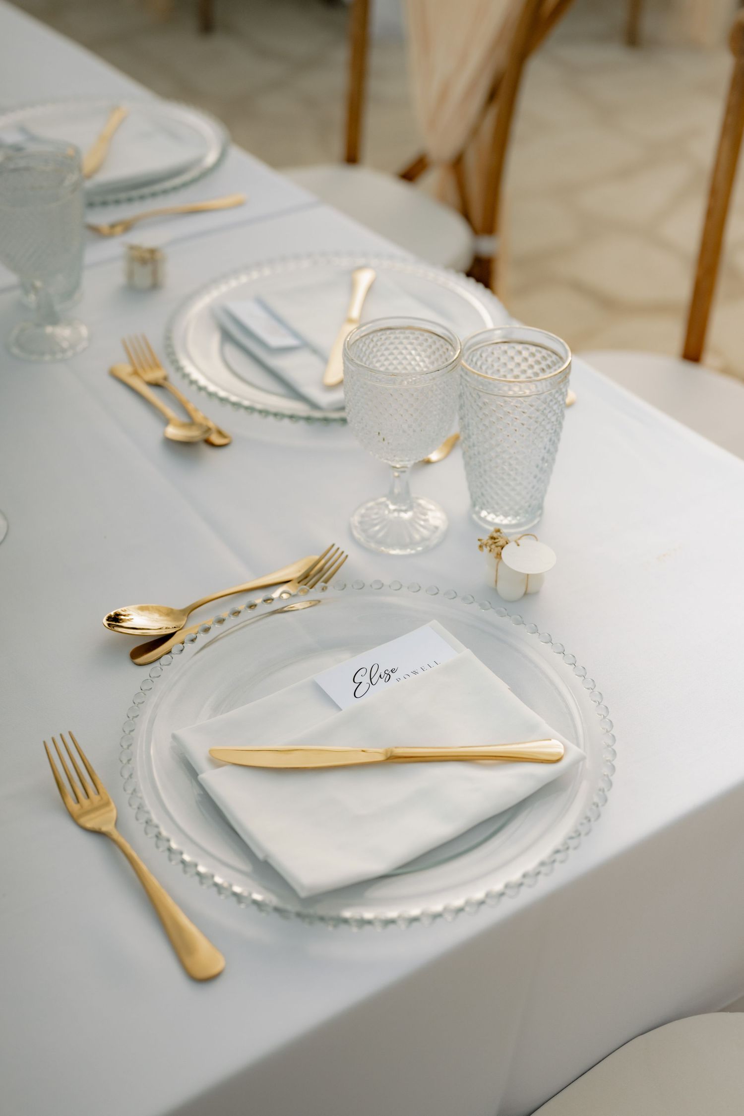Elegant wedding table setting with gold cutlery, crystal glasses, white napkins, and place cards on a gray tablecloth.