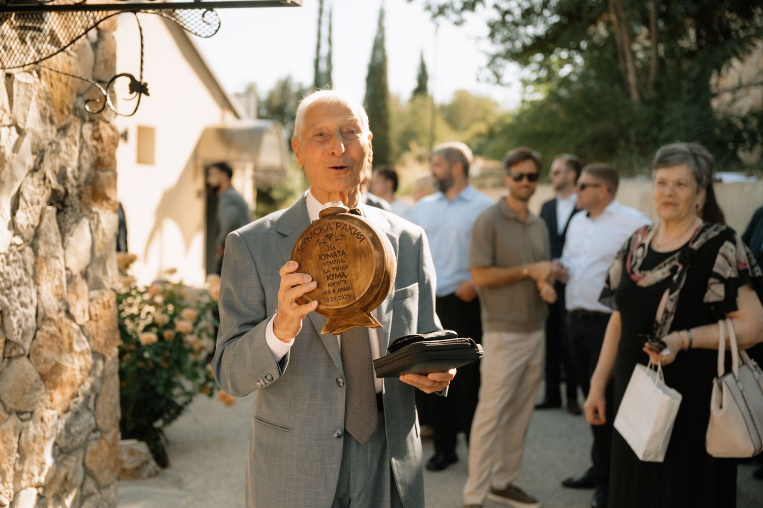 A person in a gray suit holds a ceremonial bread loaf at an outdoor gathering with guests in the background.