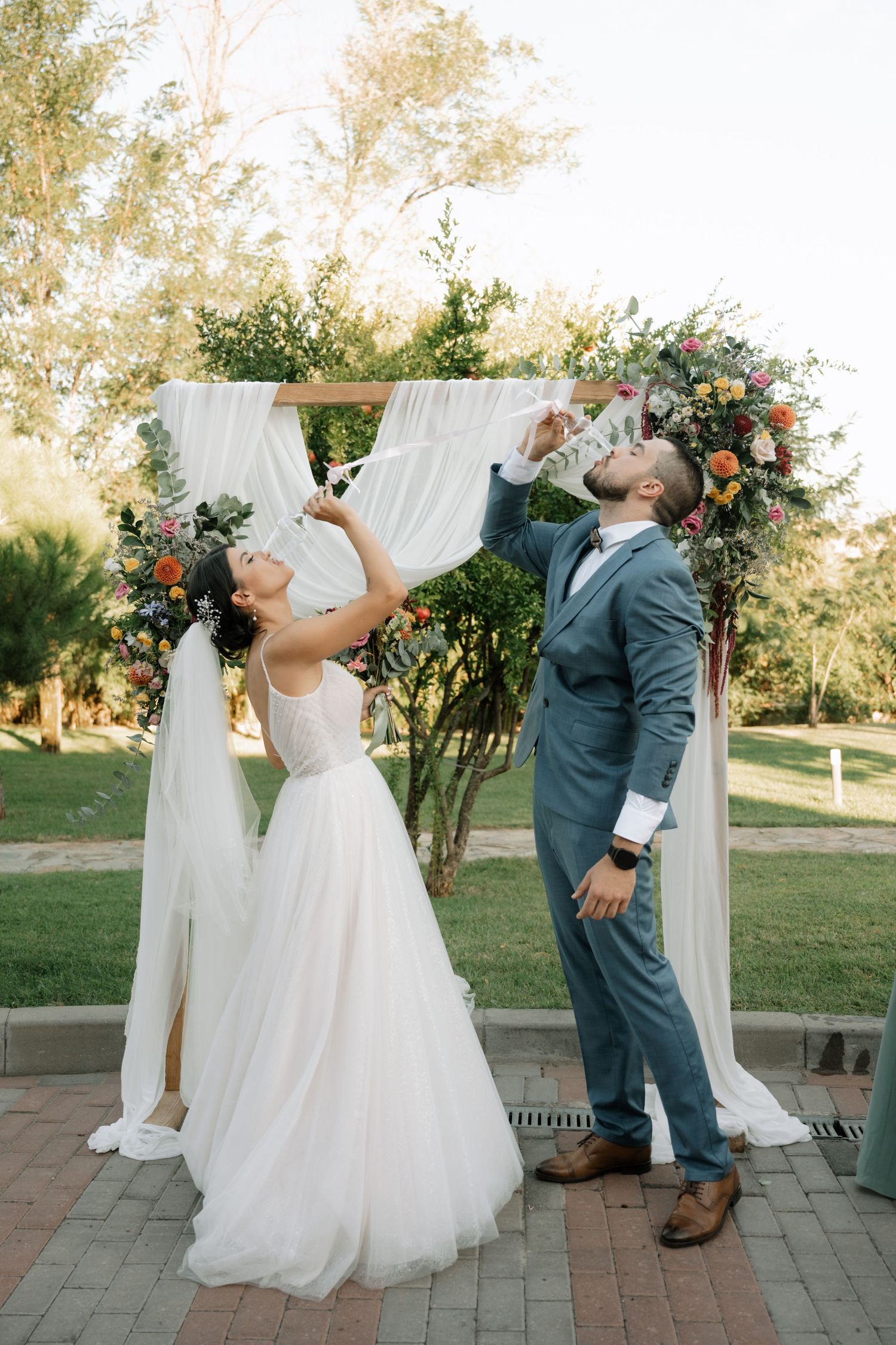 A couple celebrates under a floral wedding arch with white draping and greenery in an outdoor garden setting.