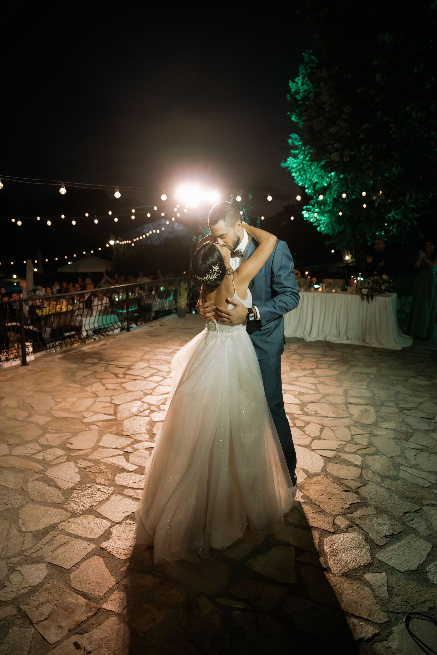 A romantic dance under string lights on a stone patio during an evening wedding reception.