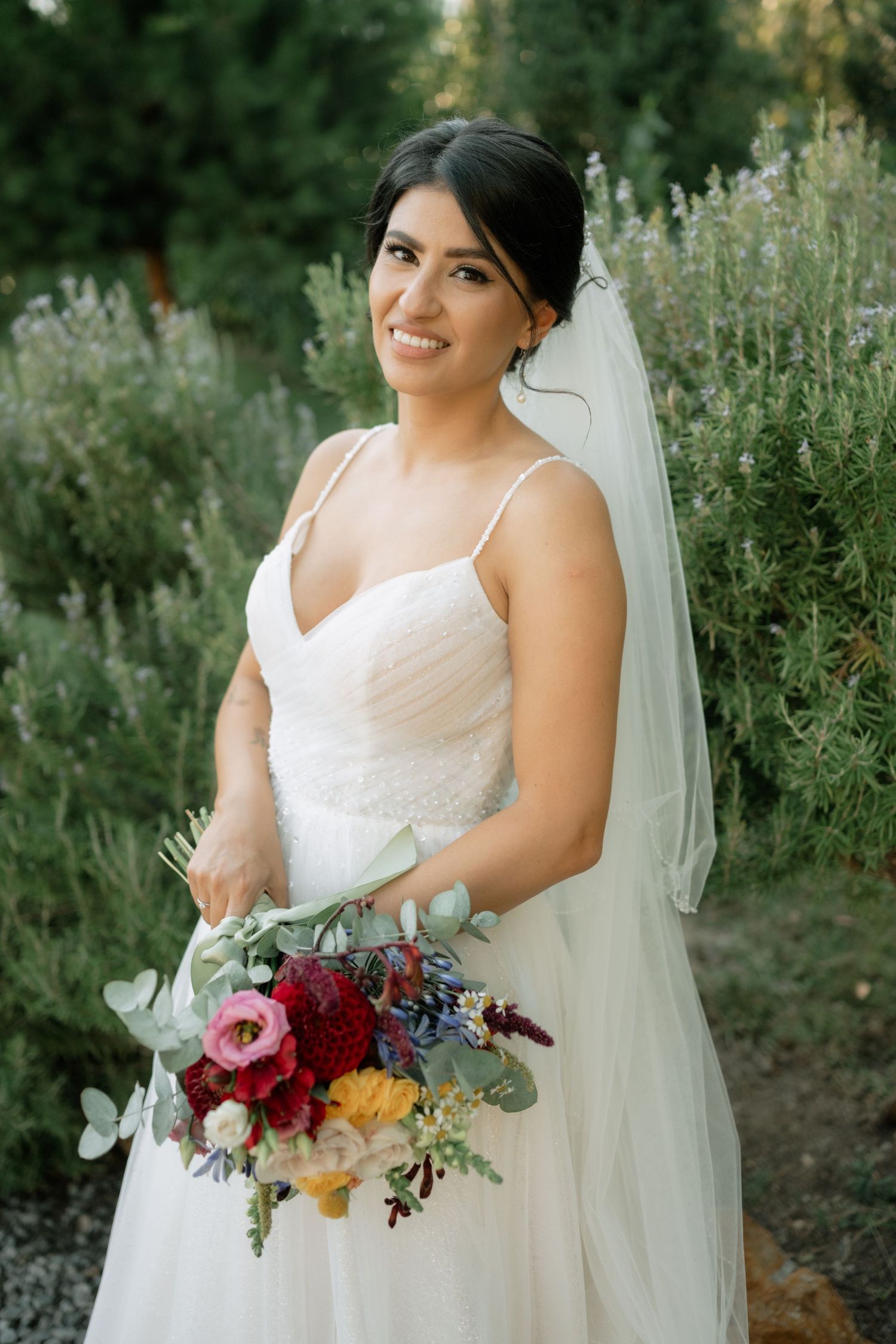 Bride in white wedding dress holds vibrant autumn-colored bouquet with burgundy and pink flowers against greenery backdrop.