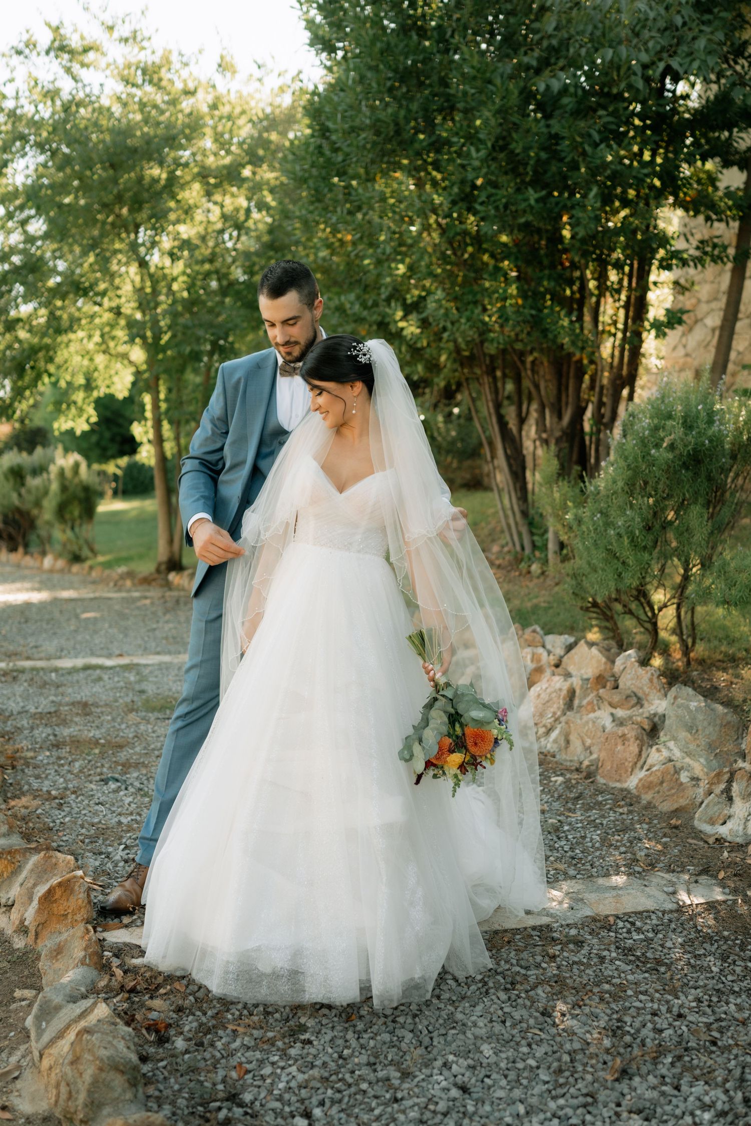 A romantic outdoor wedding moment with a couple in formal attire standing on a gravel path surrounded by greenery.