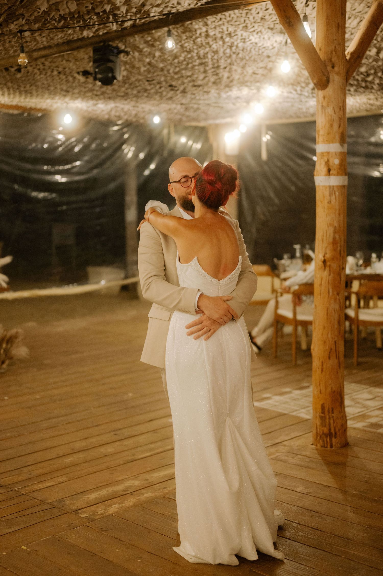 A couple shares their first dance together on a rustic wooden dance floor beneath twinkling lights at their reception.