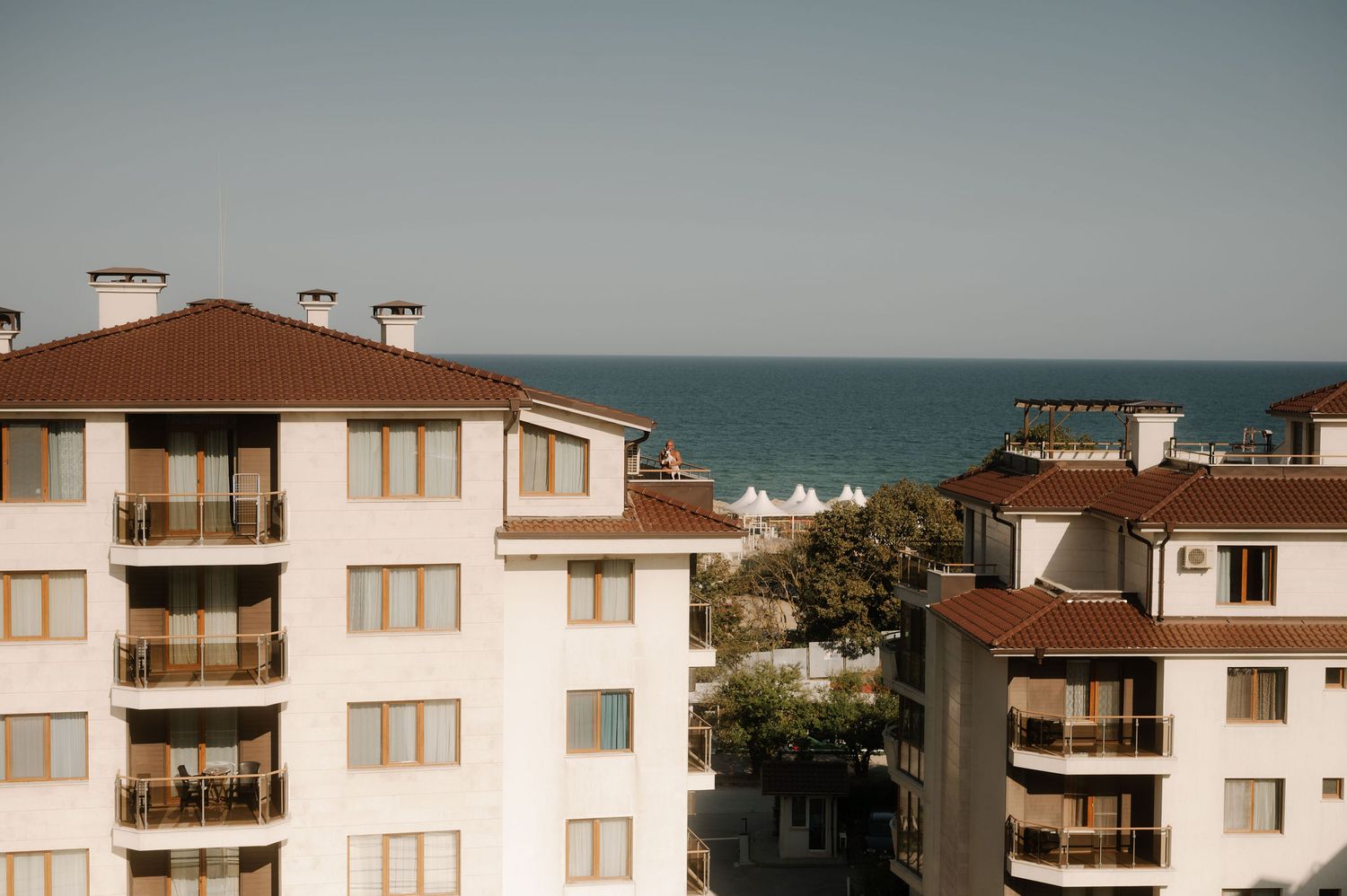 White coastal apartment buildings with brown roofs line up along the ocean horizon under an overcast sky.