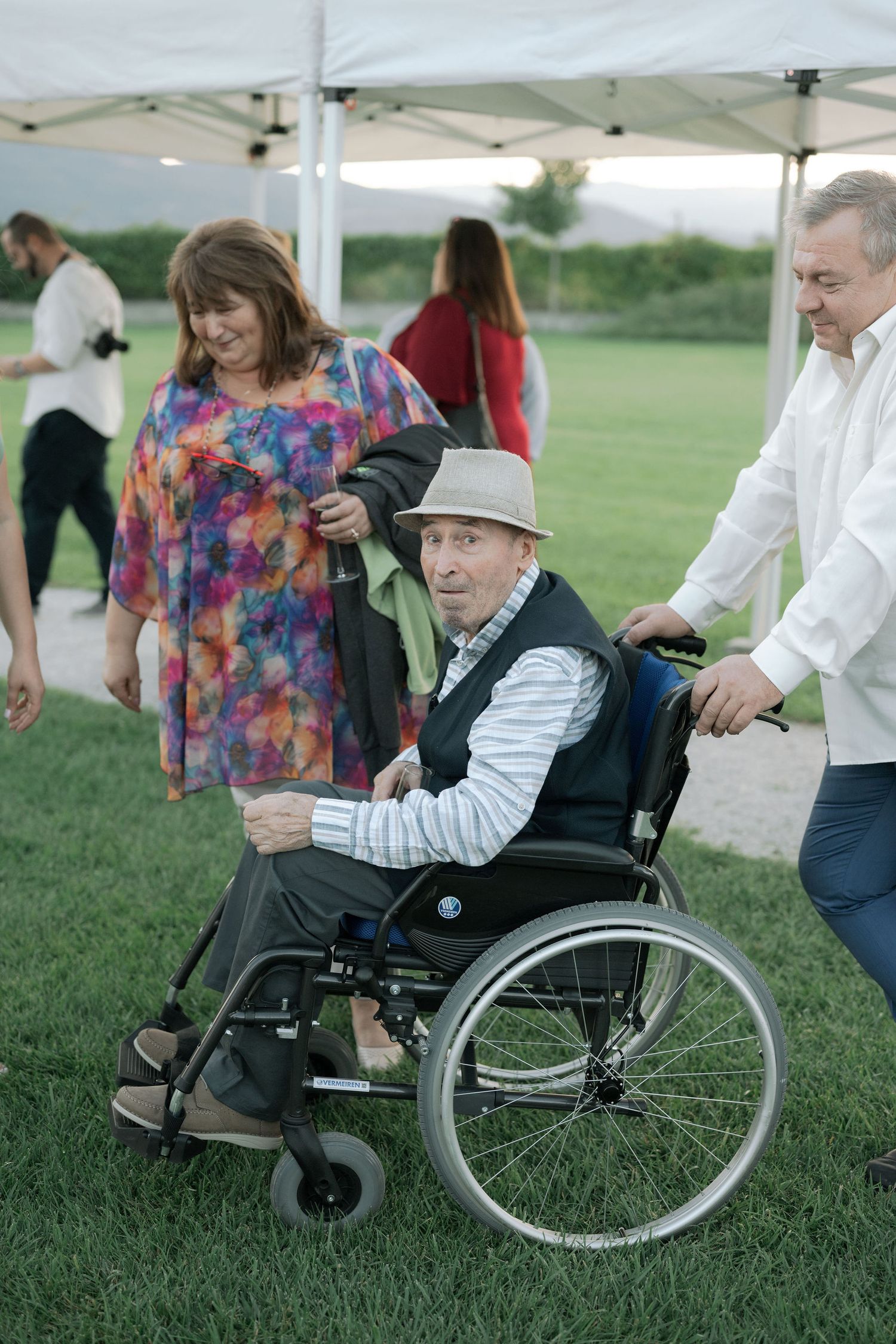 A person in a wheelchair sits on grass under a white canopy tent during an outdoor event.
